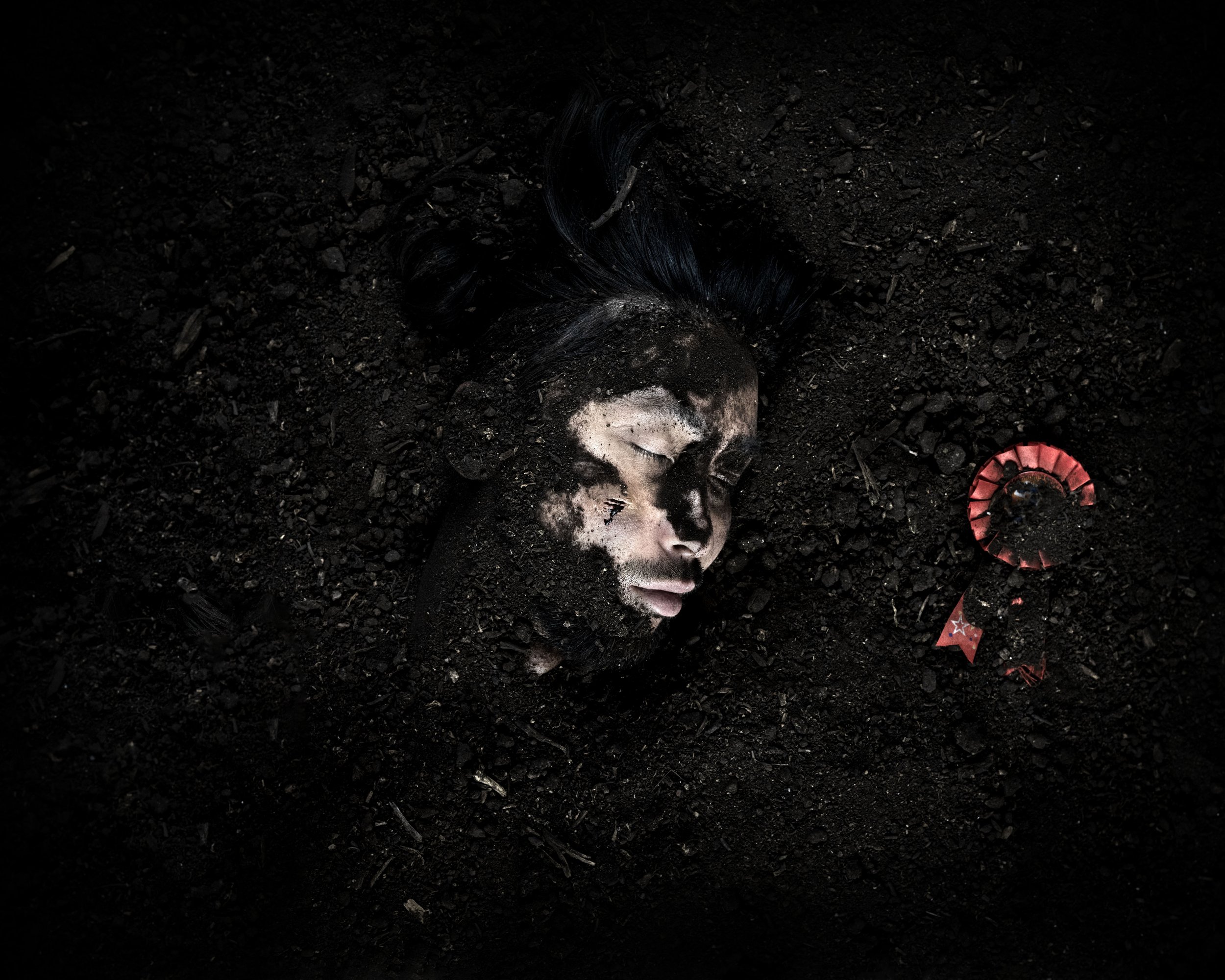 A woman with black hair and dark skin, partially covered in dirt and ash, lies peacefully on dark soil with eyes closed. Next to her is a red, broken ribbon or badge with a star on it.