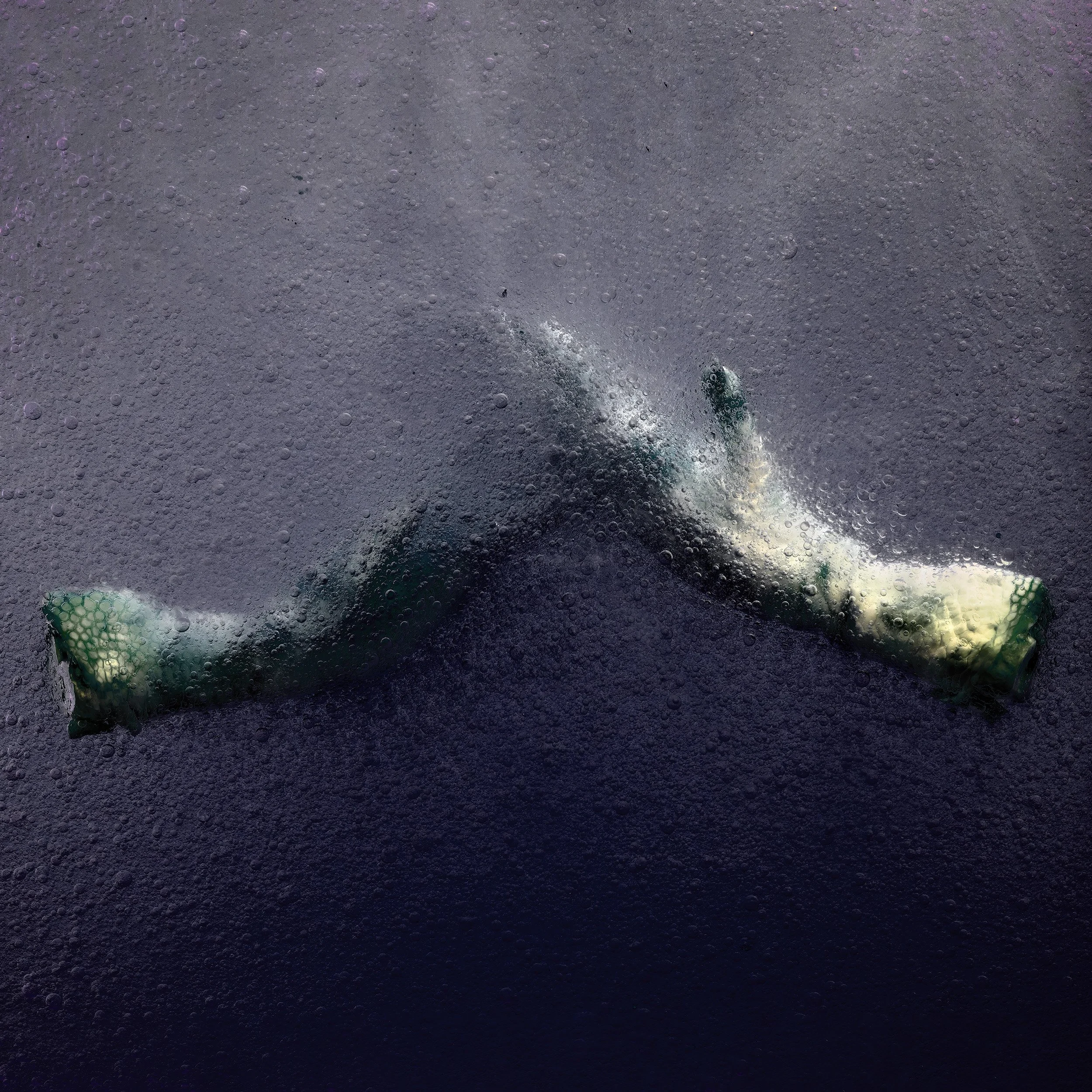 A crumpled green plastic bottle on a dark surface, with bubbles visible on the surface, suggesting the bottle is submerged in a carbonated liquid.