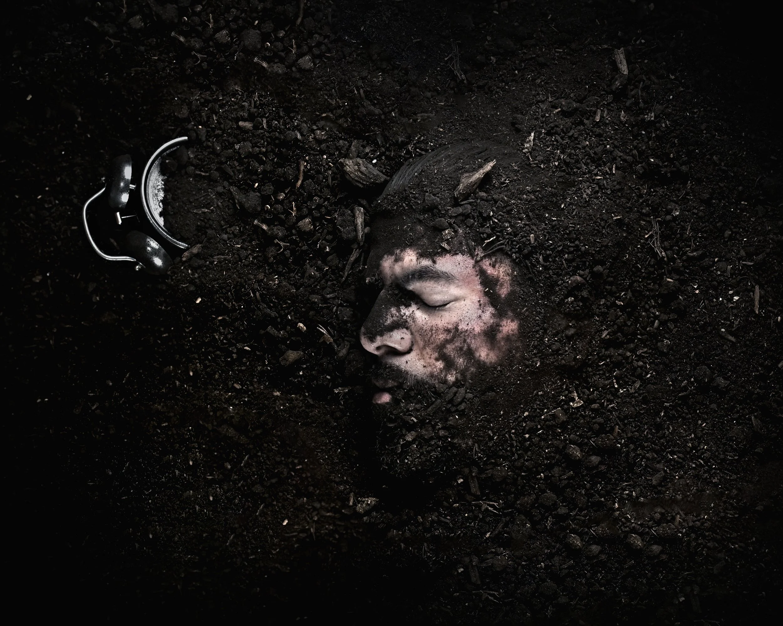 A person's face covered in dark soil, with their eyes closed, surrounded by more soil, and a black alarm clock nearby.