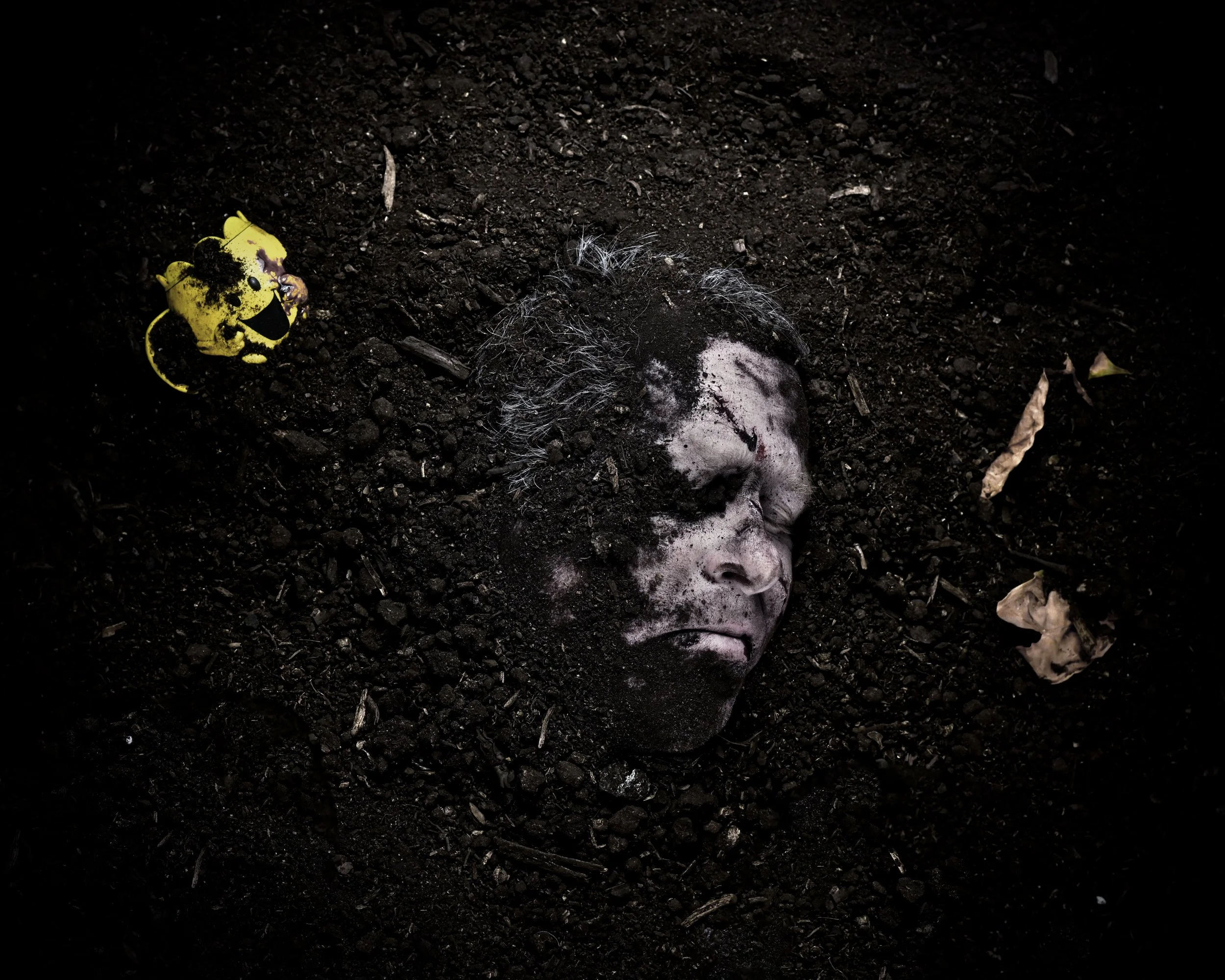 A human face with gray hair is buried in dark soil, with only the face visible, appearing dead or unconscious.