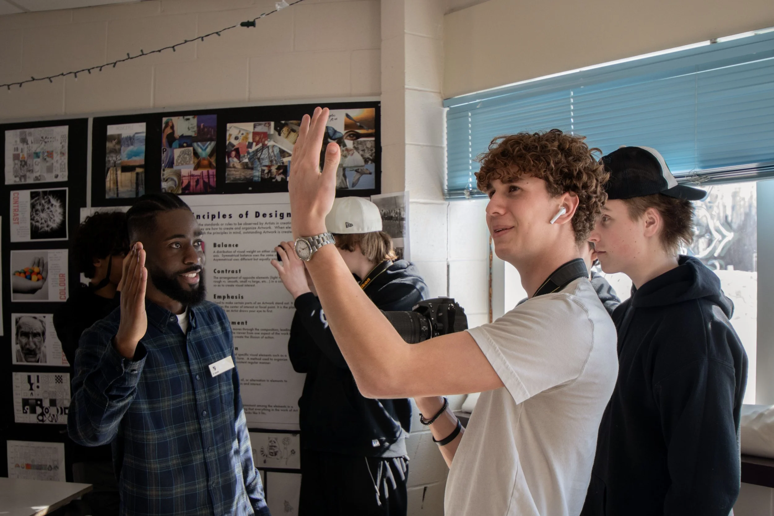 A group of young men during a tattooing or body art demonstration, with one man having a tattoo needle on his arm while others observe, and a poster about principles of design in the background.