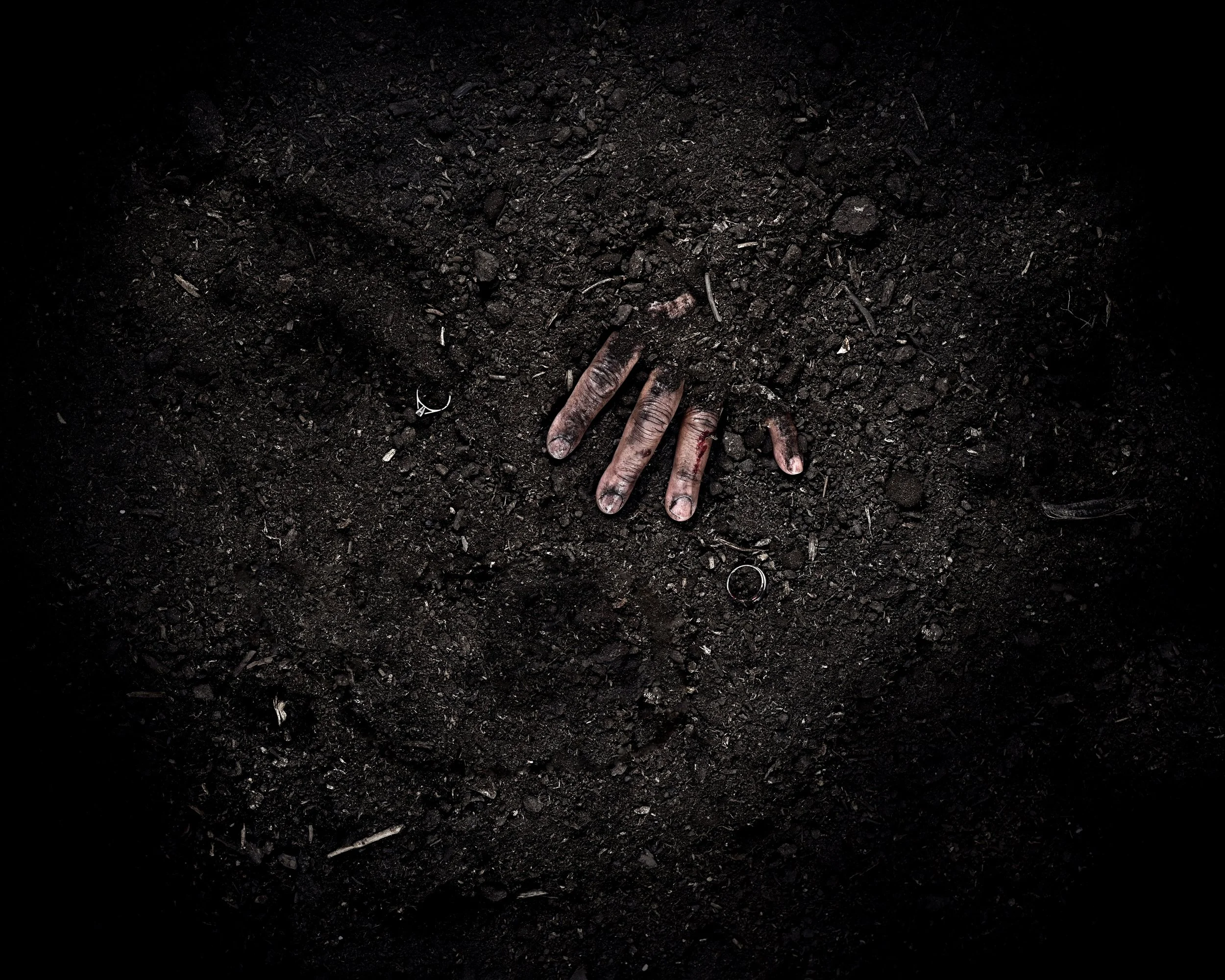 A hand with a wound and a ring lying on dark soil or dirt, surrounded by darkness.