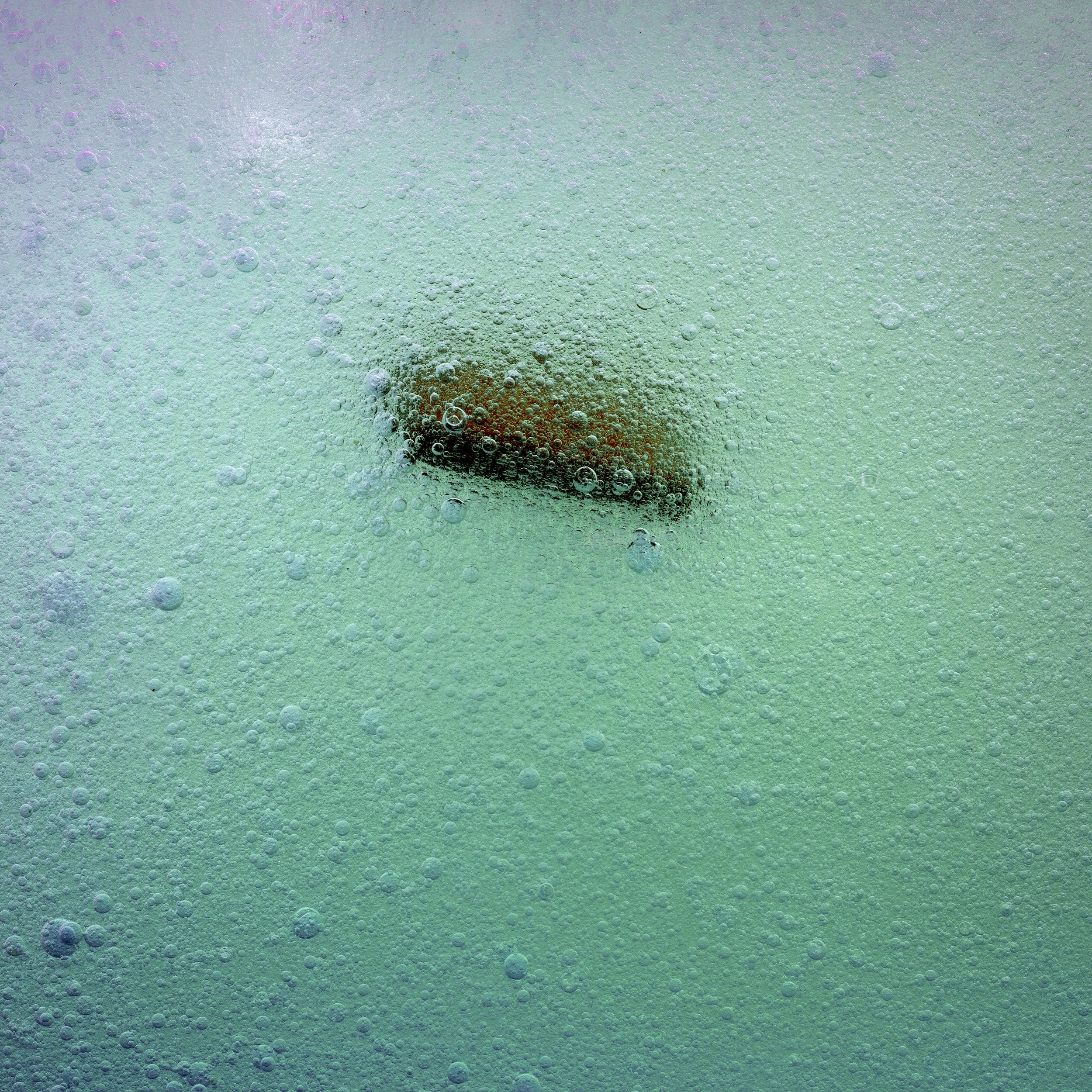 An object captured underwater in a carbonated drink, surrounded by bubbles, with a reddish-brown surface visible in the background.