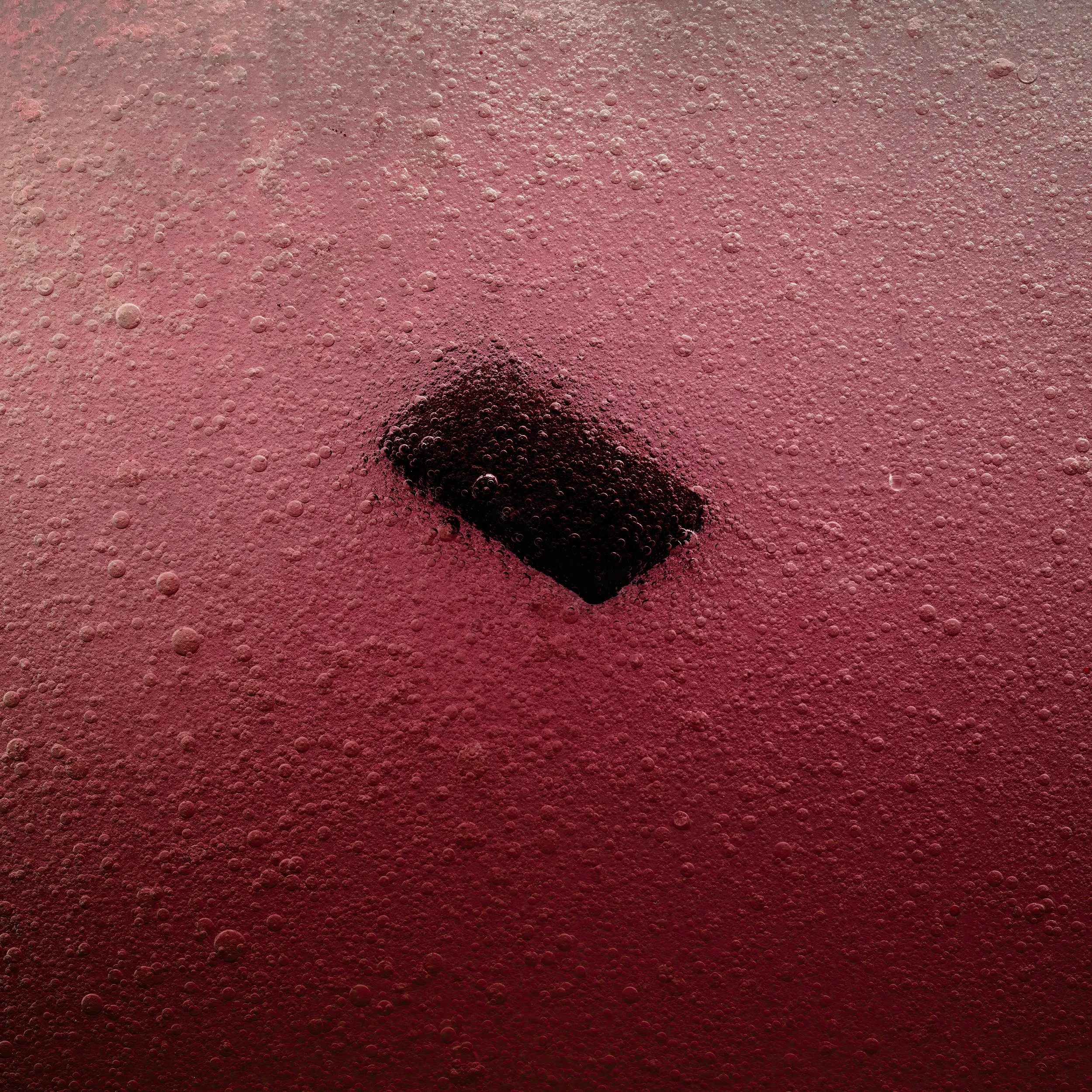 A close-up of a smartphone submerged in a pinkish-red carbonated beverage, with bubbles surrounding it.