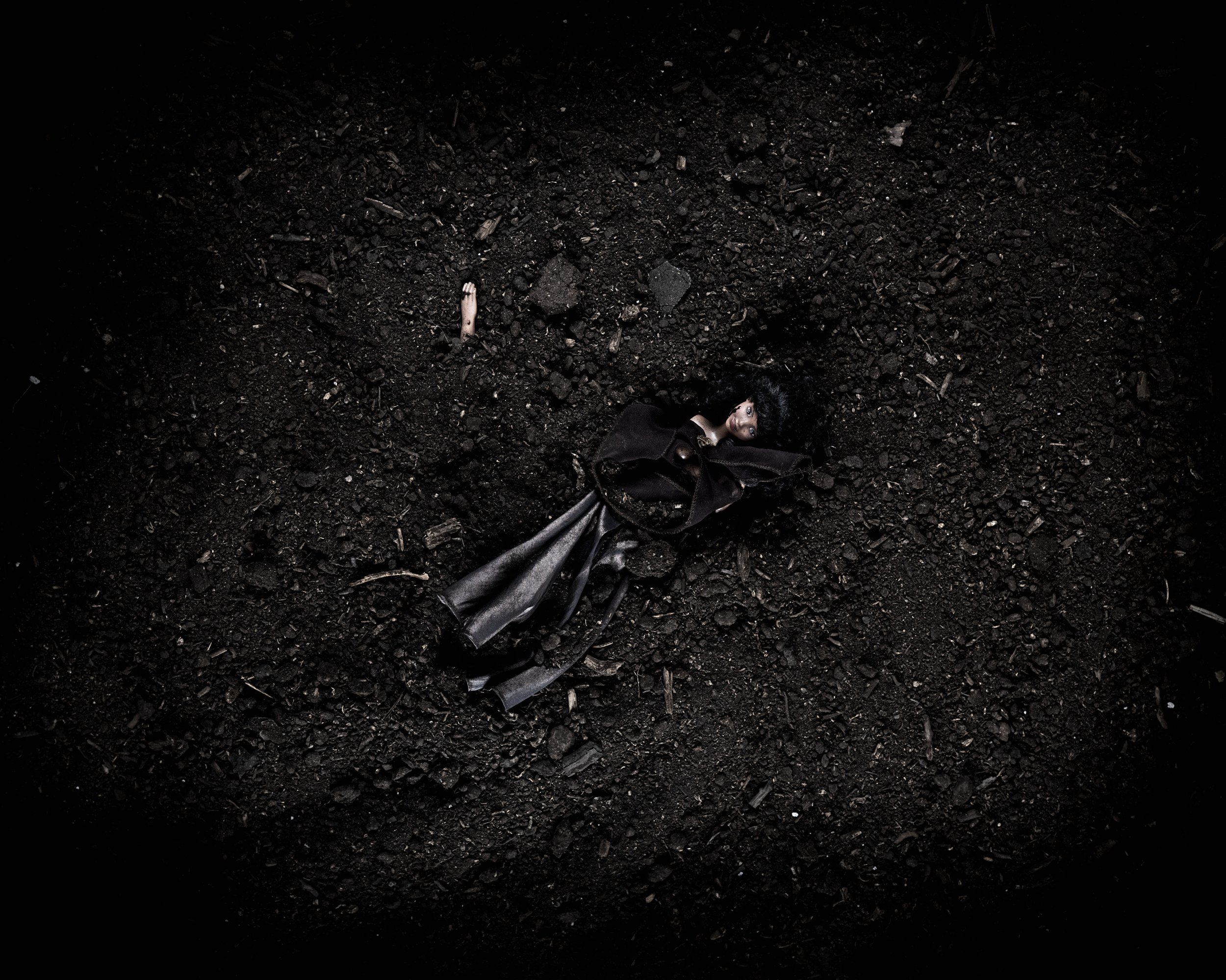 A doll with dark hair and dark clothing lying on a dark, textured surface.