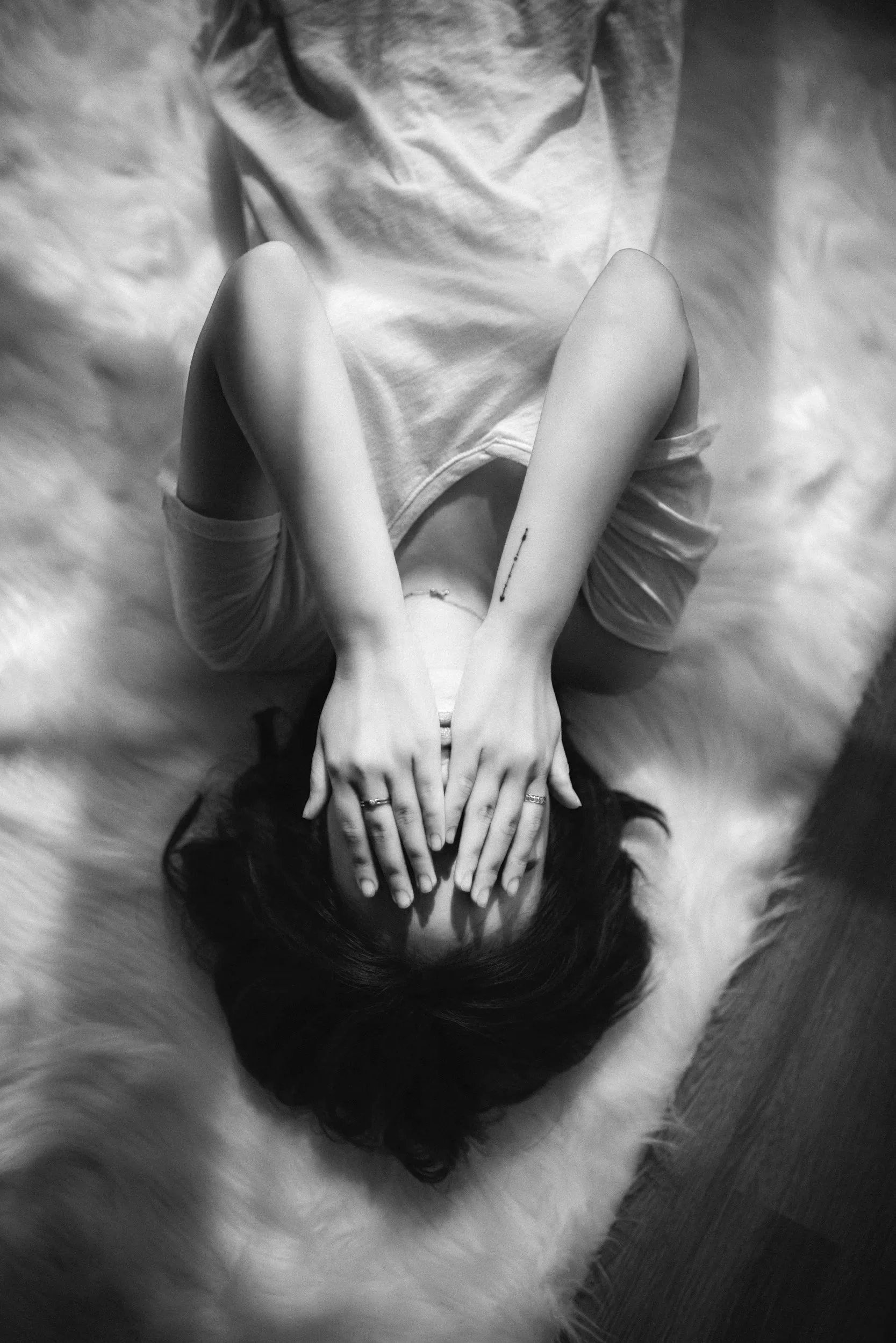 A woman lying on a fluffy rug with her hands covering her face, wearing a short-sleeved shirt and shorts, black hair, and multiple rings on her fingers.