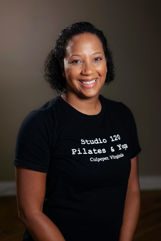 Smiling woman with curly hair wearing a black t-shirt that says 'studio 120 Pilates & Yoga Culpeper, Virginia,' standing indoors against a neutral background.