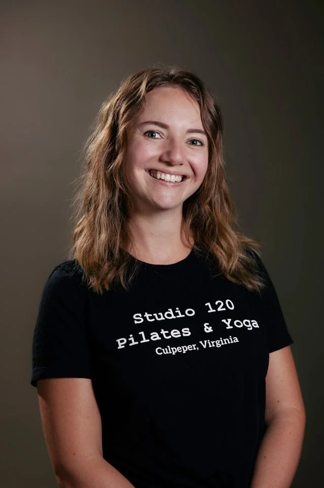 A young woman with wavy, shoulder-length brown hair, smiling, wearing a black t-shirt with white text that reads 'Studio 120 Pilates & Yoga Culpeper, Virginia'