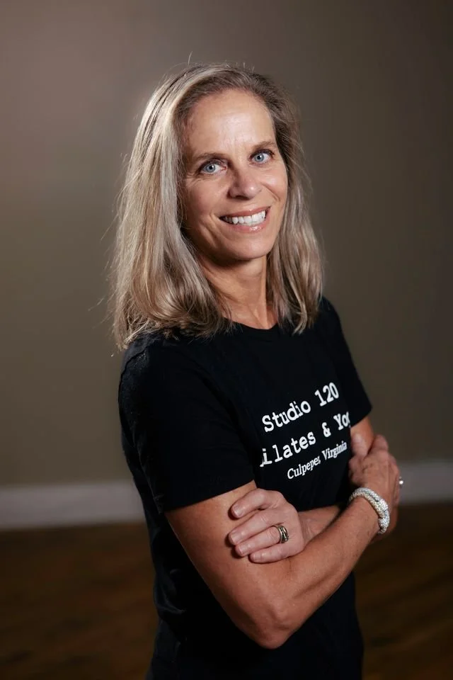 A woman with blonde hair and blue eyes standing with arms crossed, smiling, wearing a black T-shirt with text 'Studio 120 Pilates & Yoga Culpeper Virginia' and a bracelet on her left wrist.