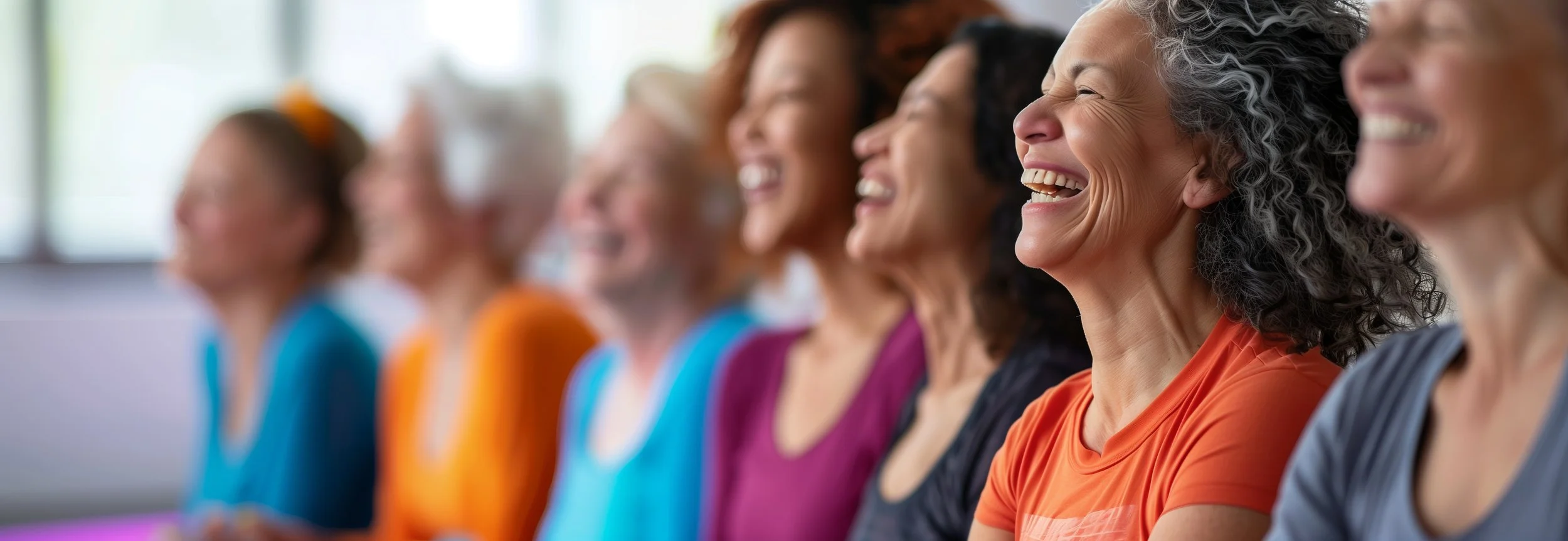 A diverse group of women sitting in a row, smiling and laughing at an indoor event.