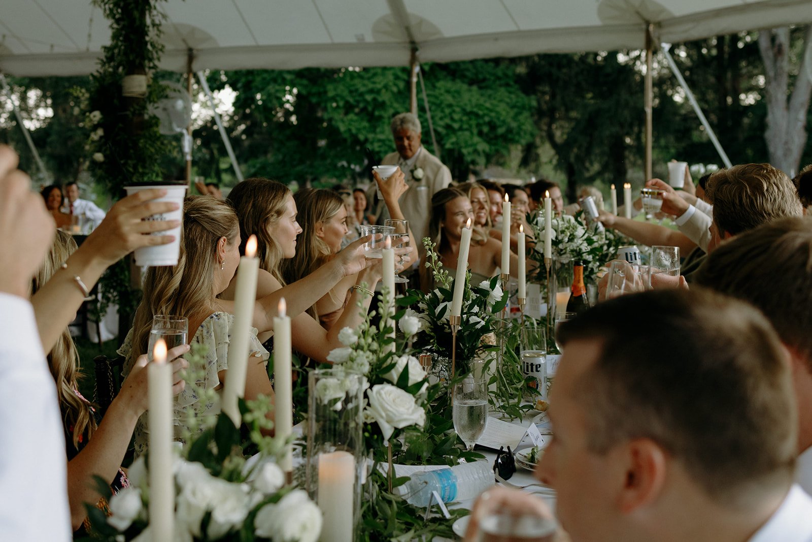 Guests raising glasses in toast at a wedding reception under a tent, with candles and flowers on the table at a classy and clean backyard wedding in Grayslake, IL by Nicole Kilday Photography