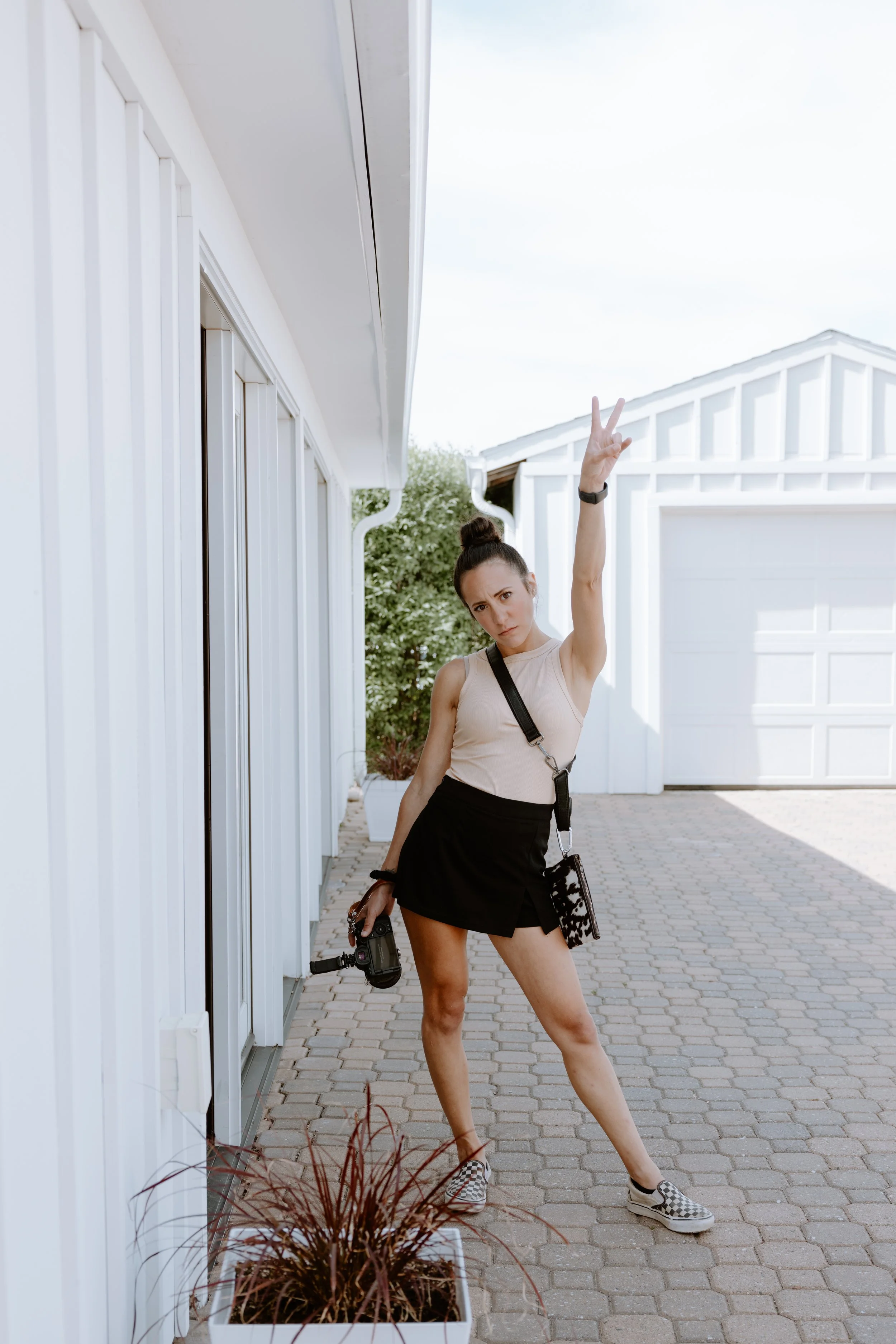 Nicole Kilday standing on a paved walkway, holding a camera in her right hand, and making a peace sign with her left hand, in front of the bridal suite at Boxed & Burlap Event Venue while photographing a wedding