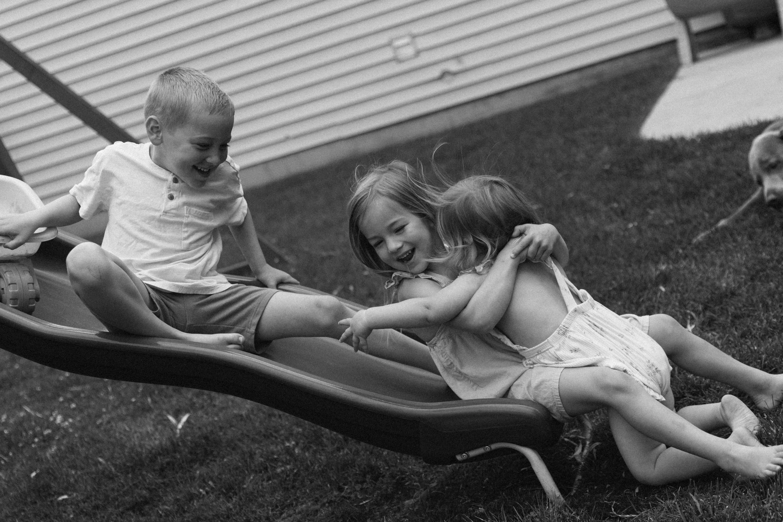Three kids laughing and playing together on a lawn chair during a lifestyle family session at home in McHenry Illinois, photographed by Nicole Kilday