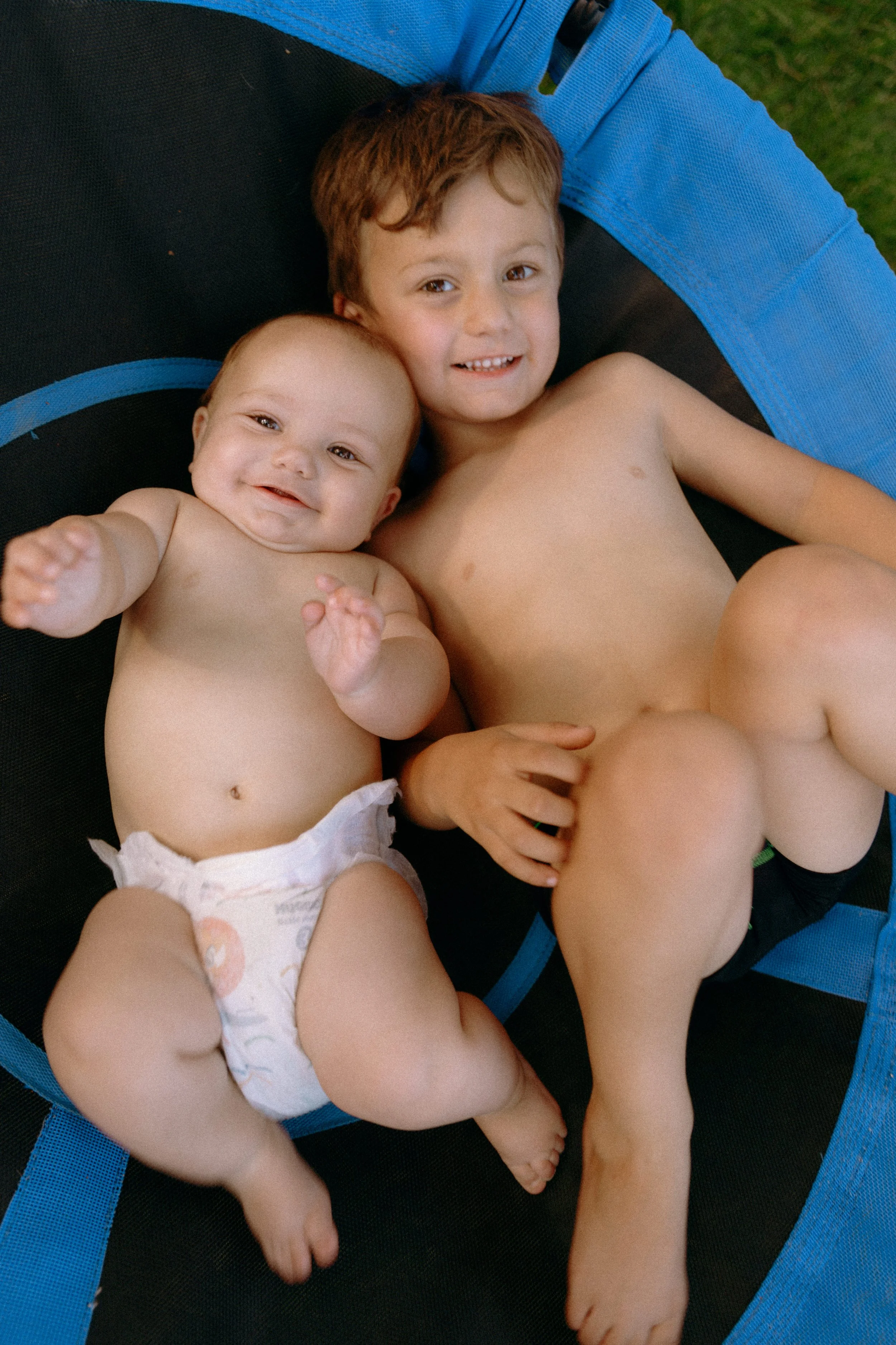 Two young children, a baby and an older boy, lying together on a trampoline, smiling and relaxed in a lifestyle family photography portrait by Nicole Kilday Photography