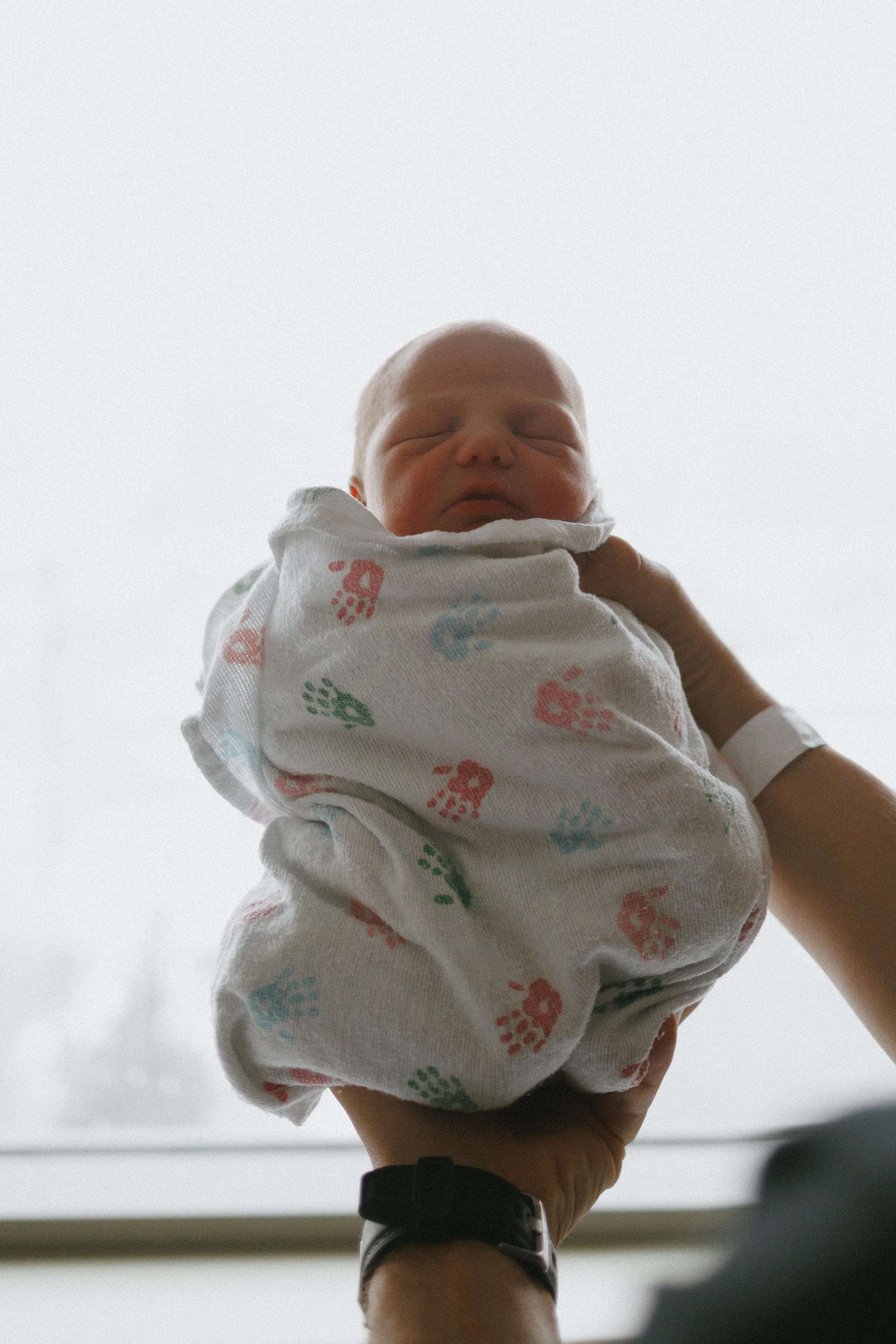 A newborn being held in front of the hospital window documenting his first day of life and the first snowfall of the year at a Fresh 48 Session