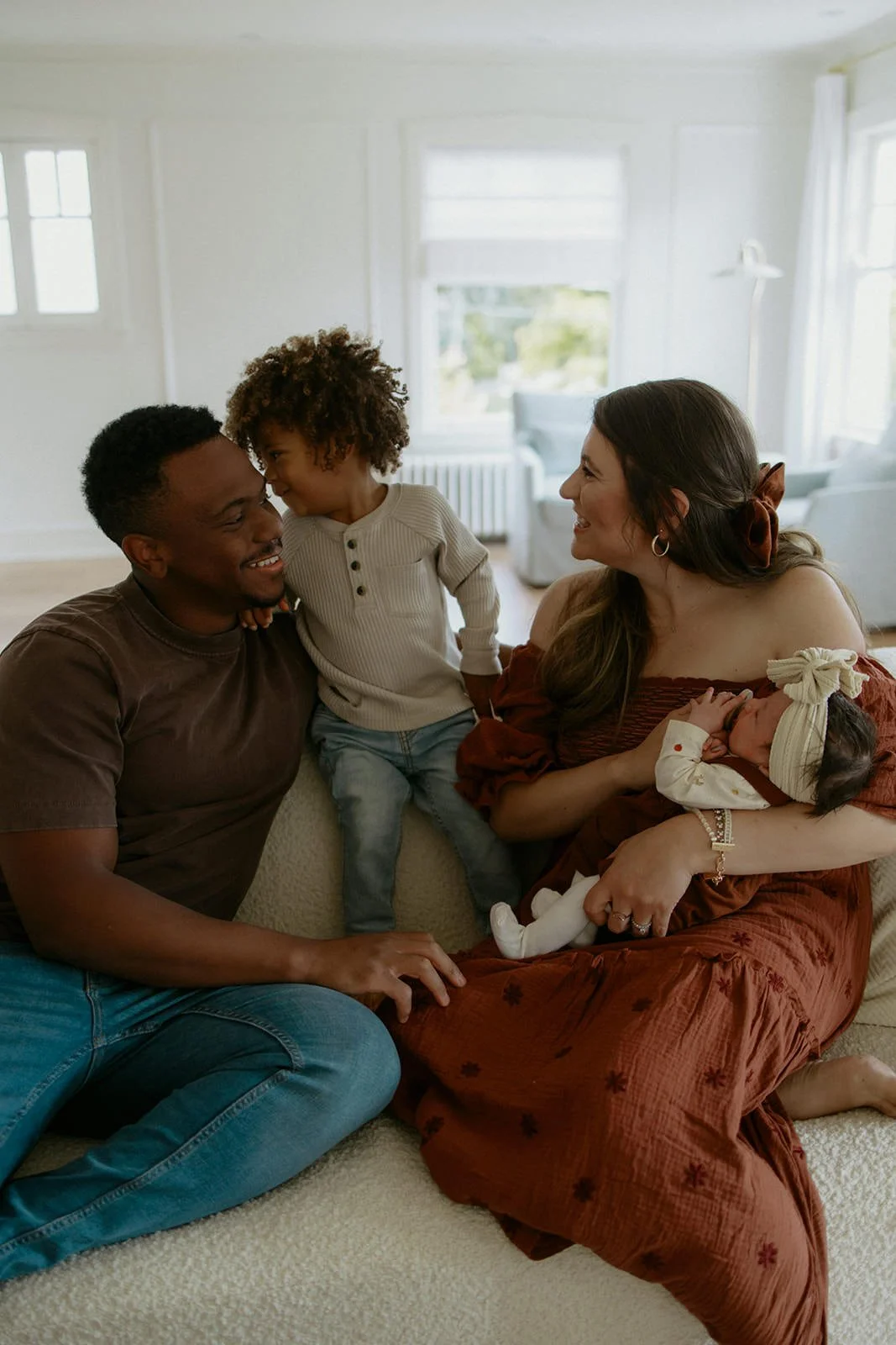 A family of four sitting on a beige couch in a brightly lit studio at The Ellery in Crystal Lake IL during their newborn family photo session indoors in the fall with Nicole Kilday Photography