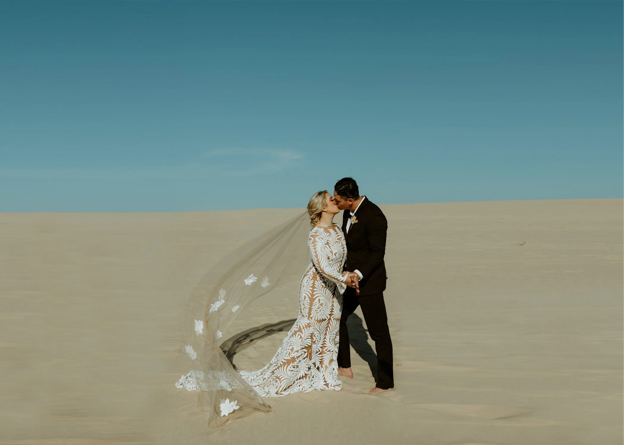 Newlyweds sharing their first kiss on the Silver Lake Sand Dunes in Michigan, the bride's cathedral veil billowing in the wind against an open blue sky — Silver Lake Sand Dunes elopement photographed by Nicole Kilday