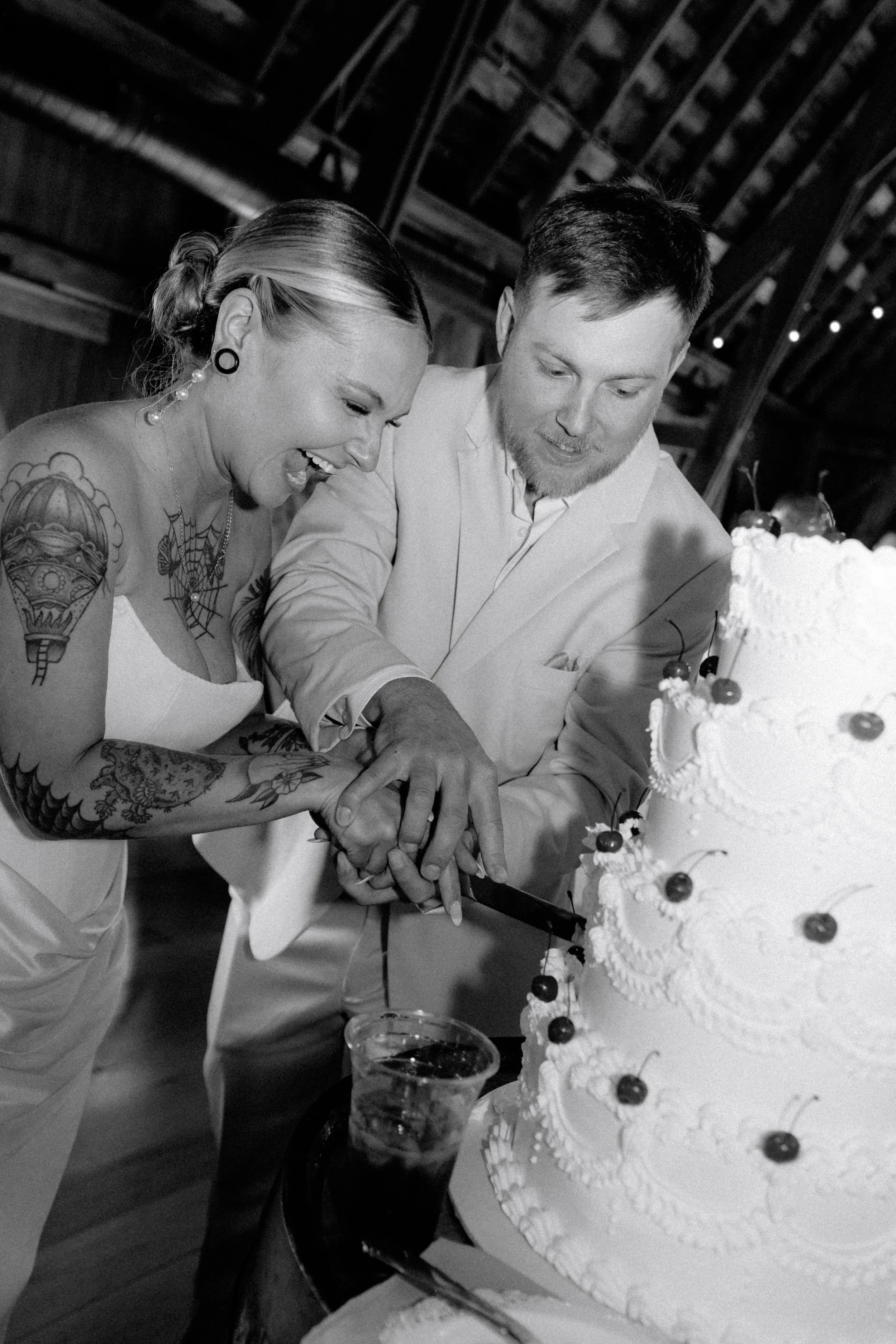 A black and white photo of a bride and groom cutting their wedding cake together, smiling and holding a knife, with the bride showing tattoos on her arm.
