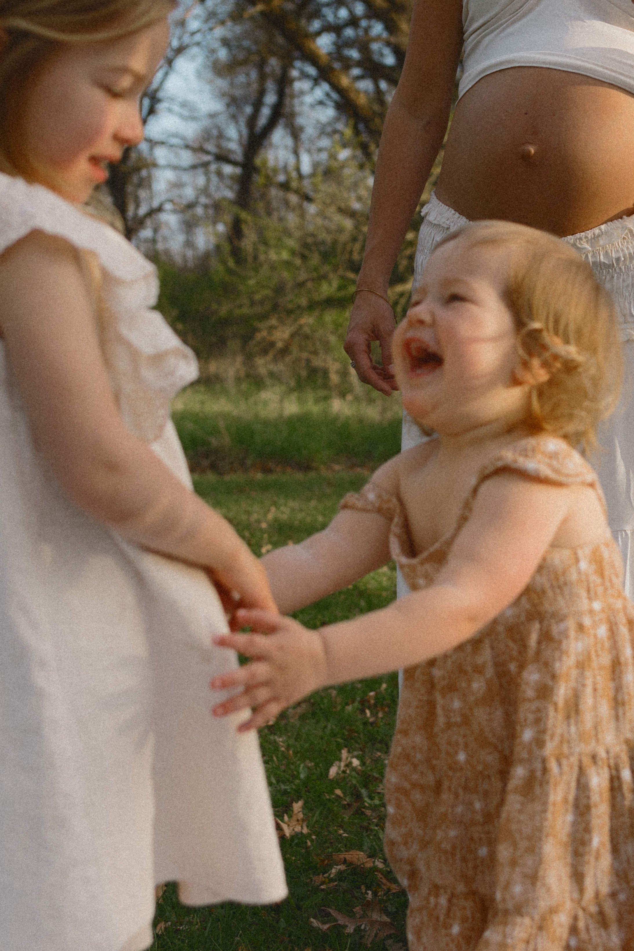 Toddler holding hands with her big sister while her  pregnant mom watches during an outdoor maternity and newborn family session in McHenry County, Illinois.