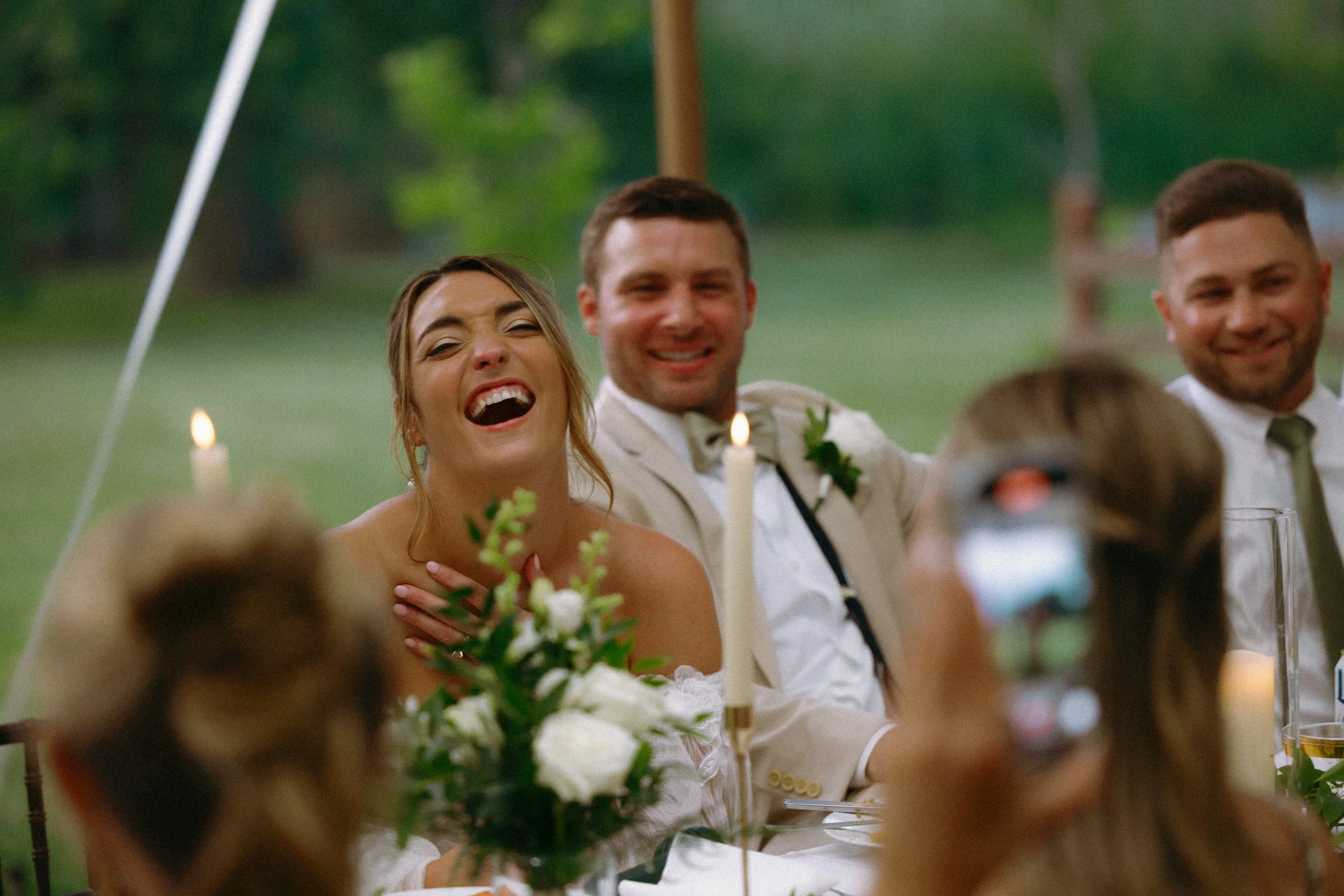 Bride laughing at the head table during wedding reception toasts, groom smiling beside her in a candlelit outdoor tent — Illinois and Wisconsin wedding photography packages, Nicole Kilday
