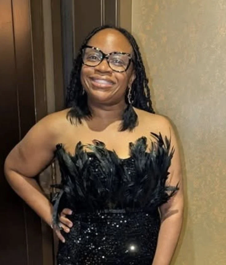 Smiling woman in glasses wearing a black feathered dress at an indoor event.