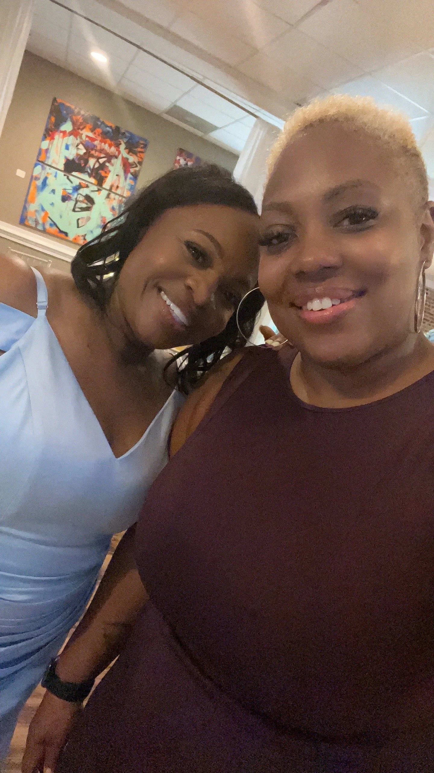Two women smiling for a selfie, one with black hair and the other with blonde hair, indoors with colorful abstract artwork on the wall behind them.