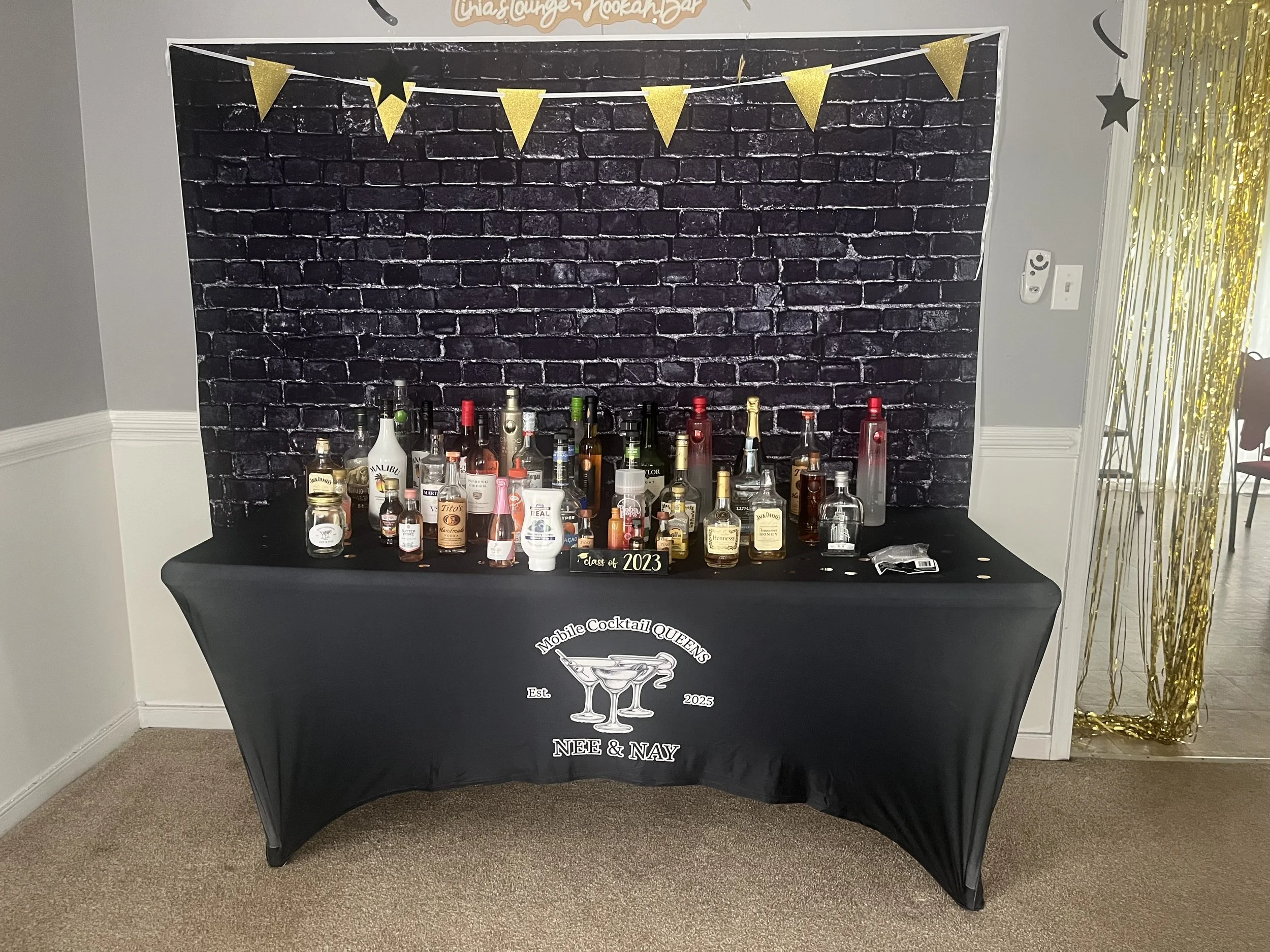 Table with various bottles of alcohol and mixers, featuring a black tablecloth with a logo that reads "Mobile Cocktail Queens NEF & NAY." Behind the table is a black brick wall backdrop decorated with a gold pennant banner and some black and gold star cutouts.