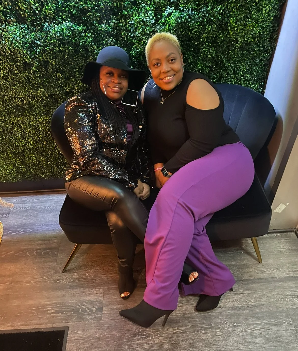 Two women sitting on a black chair in front of a green leafy wall. One woman wears a black hat, a sequined black jacket, and black leather pants. The other woman has short blonde hair, wears a black top with cut-out shoulders and purple pants. Both women are smiling.