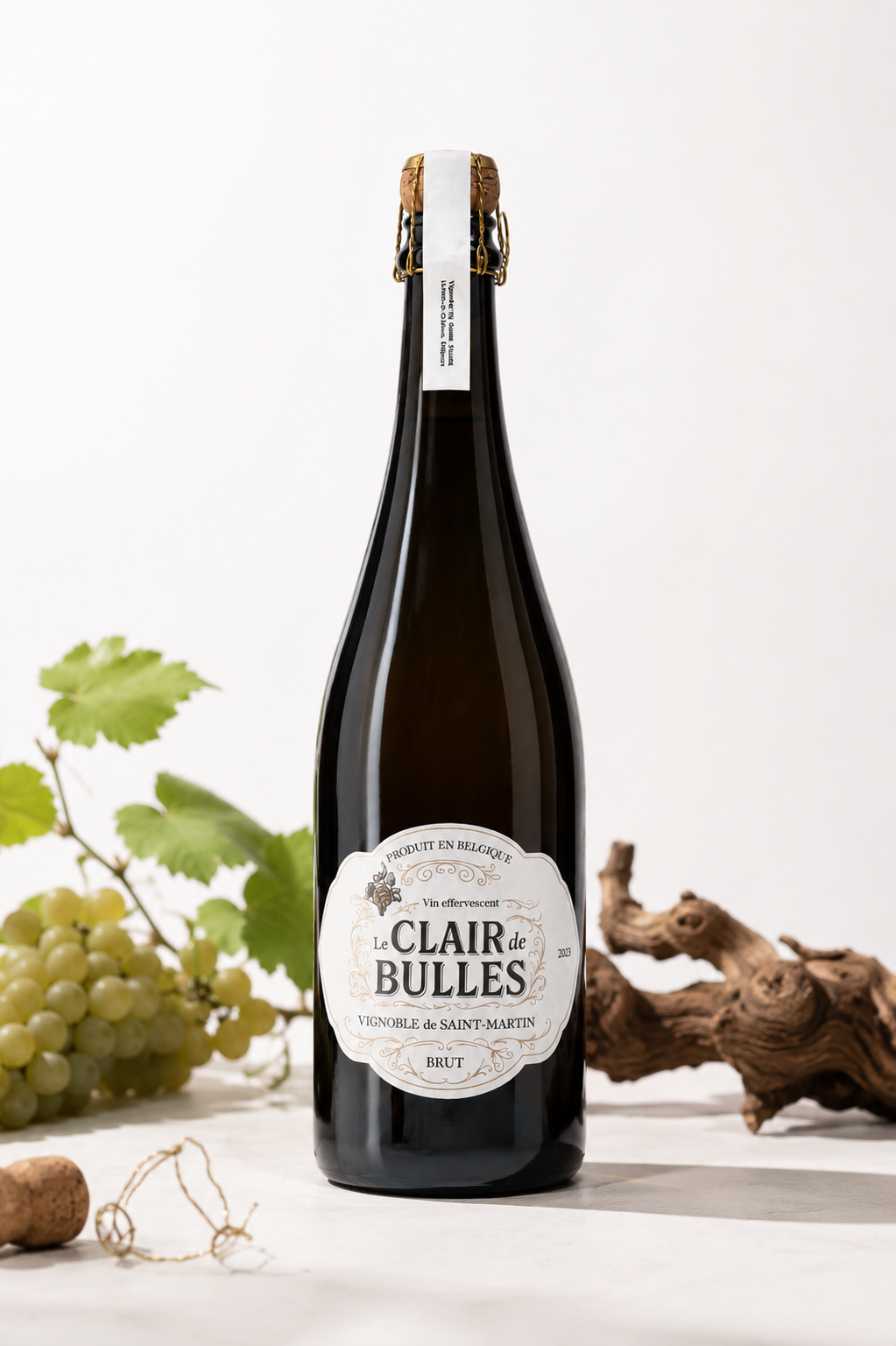 Bouteille de vin mousseux Le Clair de Bulles, entourée de raisins, de bois et de feuilles de vigne sur une surface blanche.