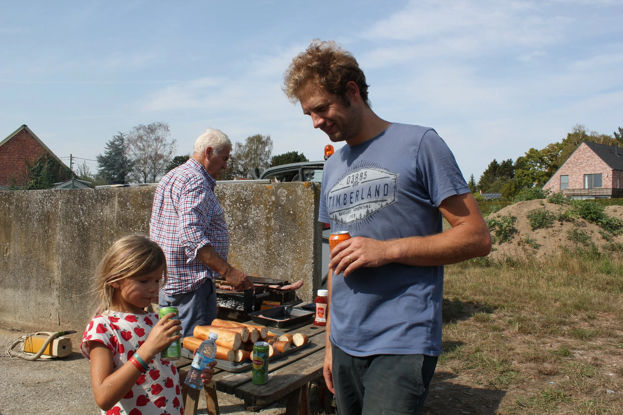Groupe de personnes autour d'une table en plein air, avec des enfants et un adulte, partageant un repas avec des saucisses grillées et des boissons, dans un environnement résidentiel.