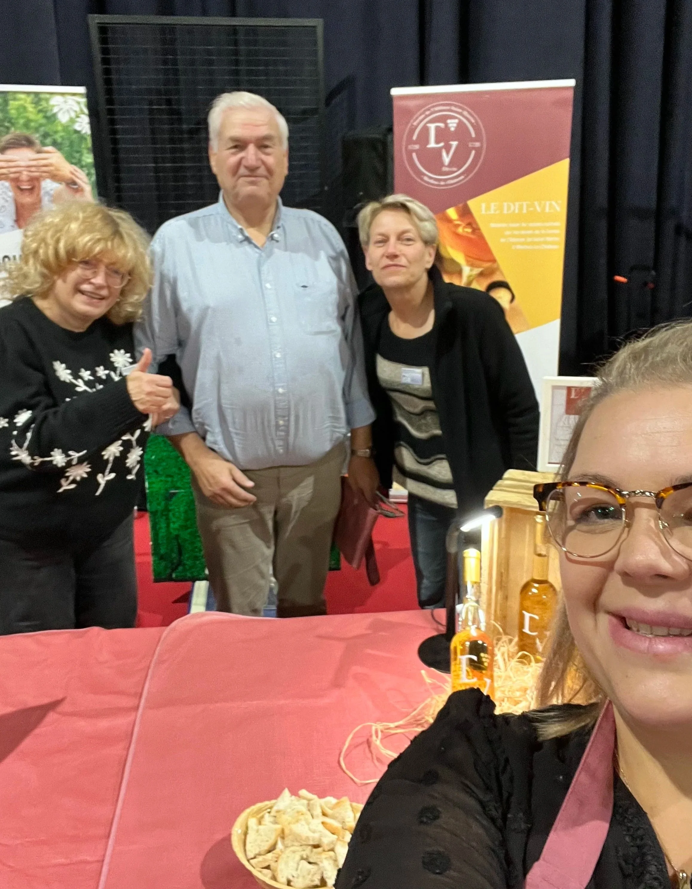 Groupe de personnes posant pour une photo lors d'un événement, avec un décor d'affiches et de bouteilles sur une table en premier plan.