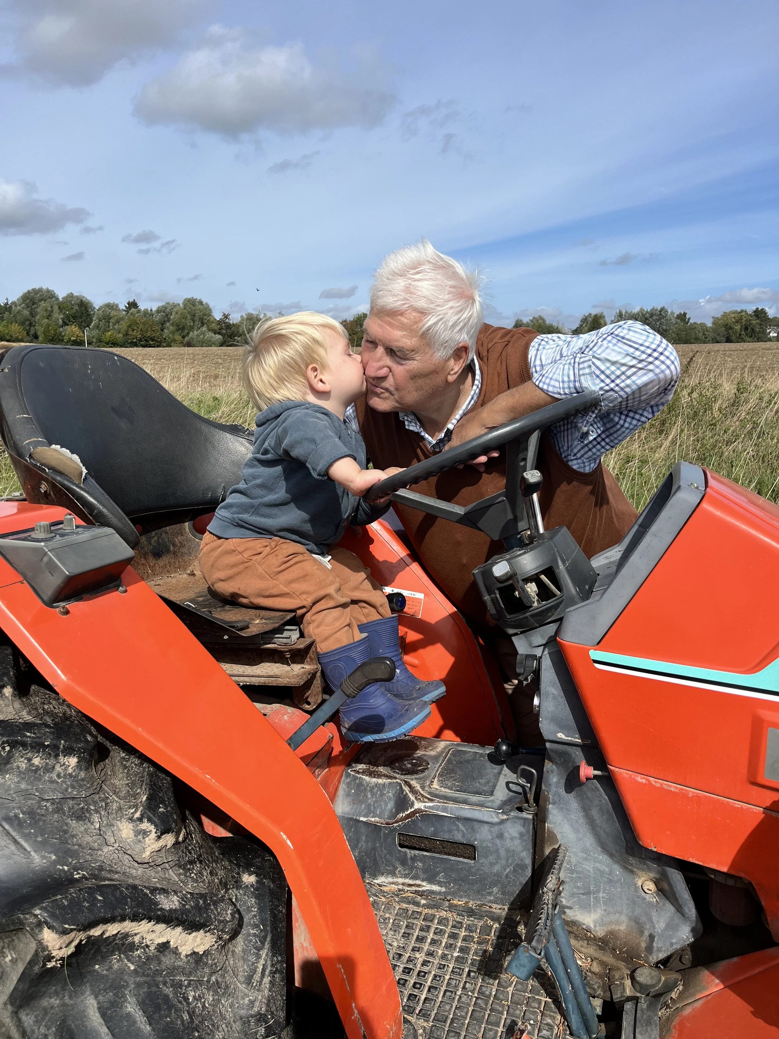 Un homme âgé embrasse tendrement un jeune garçon dans un champ ouvert avec un tracteur rouge, un ciel bleu et quelques nuages dans le fond.