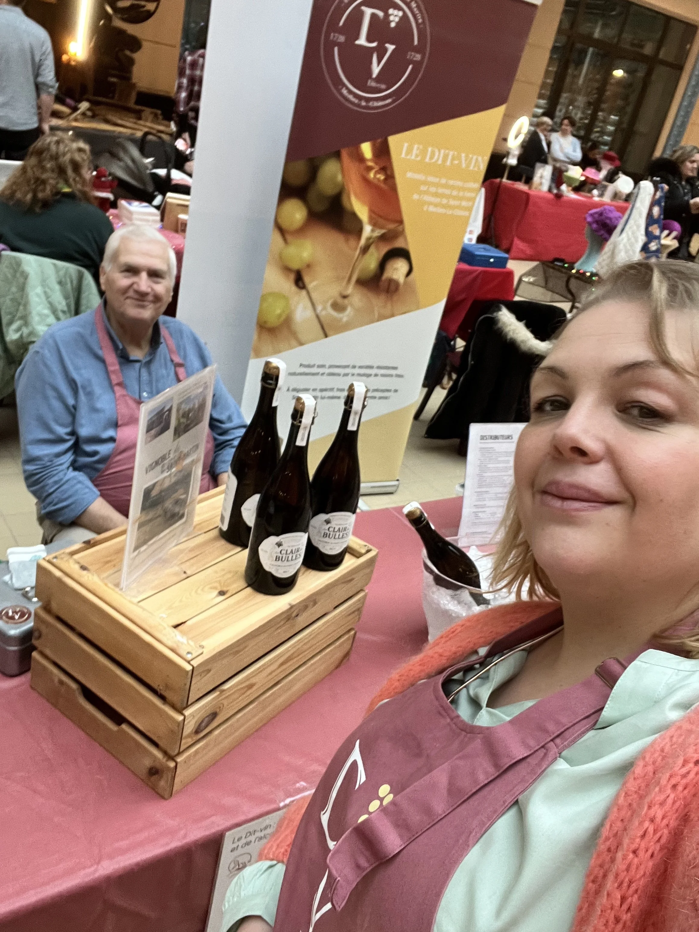 Une femme prend un selfie lors d'un événement où elle est assise à une table avec une affiche publicitaire pour des bouteilles. Il y a d'autres personnes en arrière-plan dans une salle chaleureuse.
