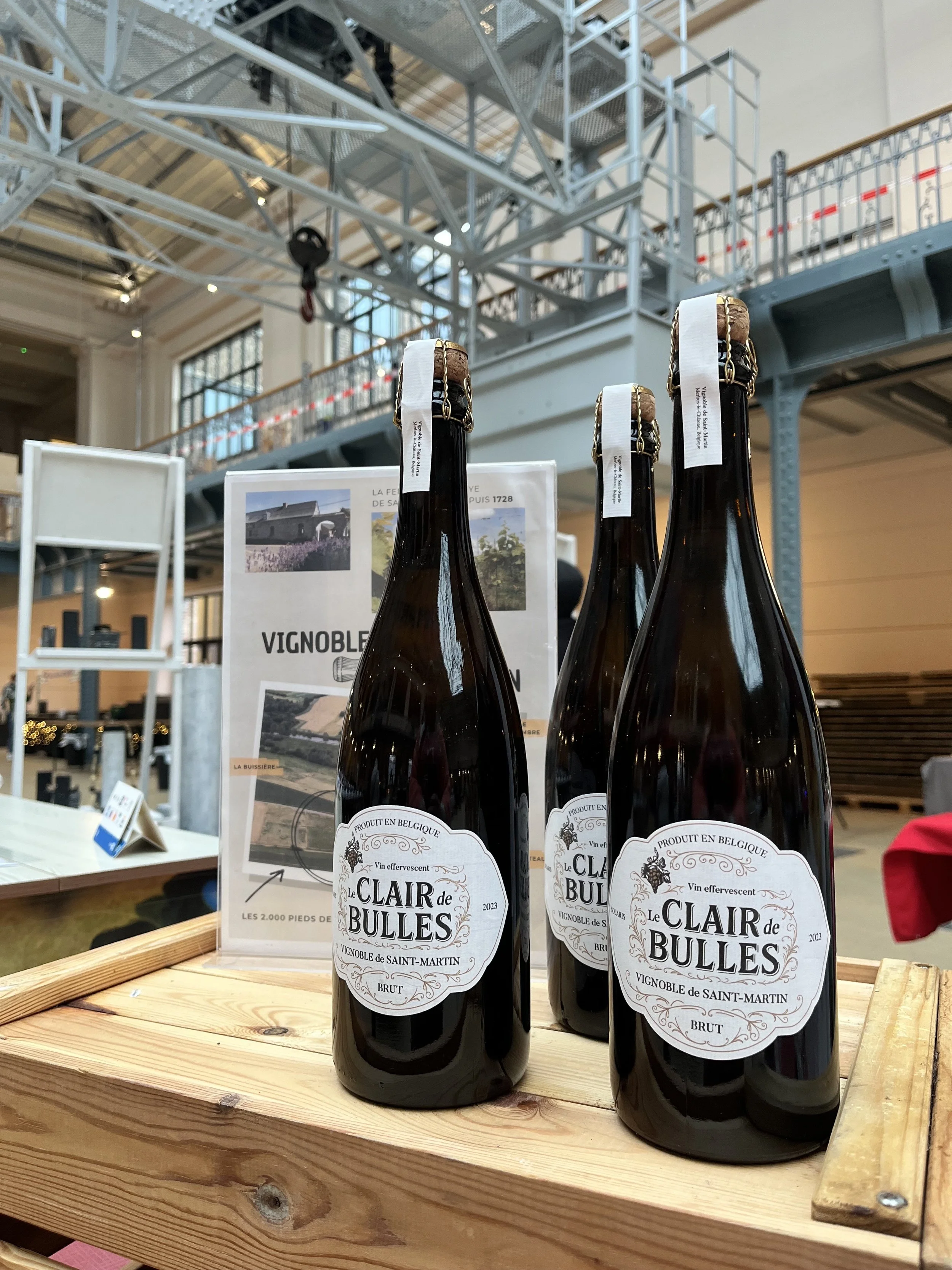 Trois bouteilles de vin effervescent nommées 'Le Clair de Bulles' posées sur une table en bois dans un espace intérieur, avec un panneau d'information derrière elles.
