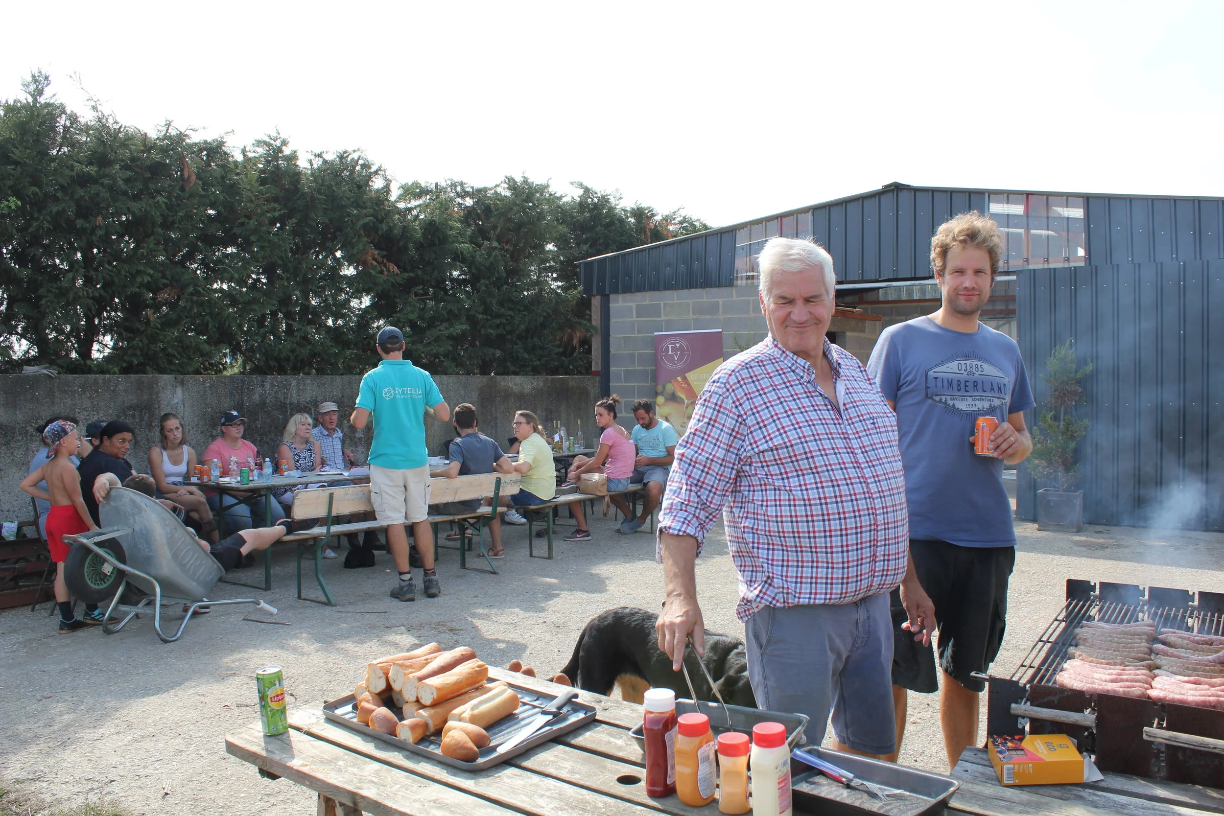 Un barbecue extérieur lors d'un rassemblement familial ou entre amis avec plusieurs personnes assises autour d'une table. Deux hommes sont visibles au premier plan, l'un avec des cheveux blancs et la barbe, l'autre plus jeune avec des cheveux bouclés. Il y a de la viande en train de cuire sur le barbecue et des assaisonnements sur une table en avant-plan. En arrière-plan, des personnes discutent, certains assis à une grande table, et d'autres se tiennent debout. Il y a des arbres et un bâtiment en arrière-plan.