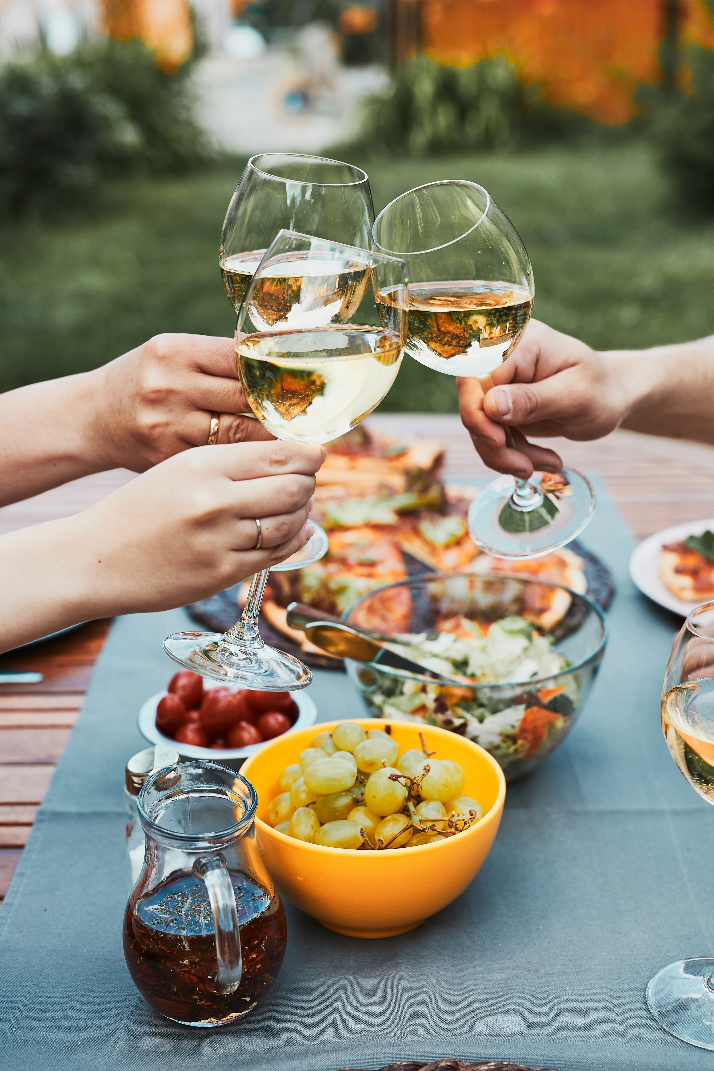 Groupe de personnes levant des verres de vin blanc pour un toast lors d'un repas en plein air, avec des plats de salade, raisins, tomates cerises et une carafe sur la table.