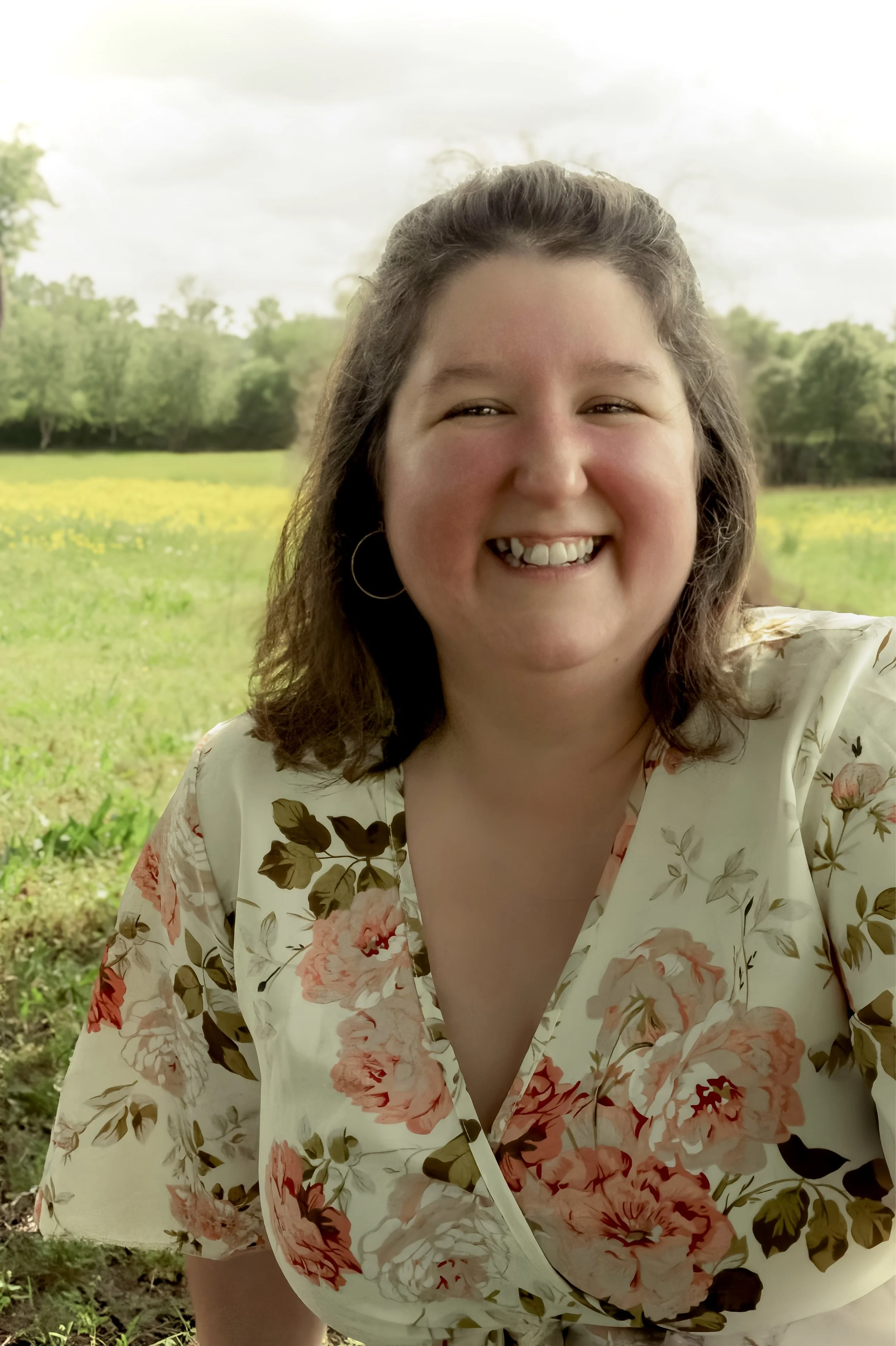 Smiling woman with shoulder-length brown hair, wearing hoop earrings and a floral blouse, outdoors in a green field with trees and a cloudy sky in the background.
