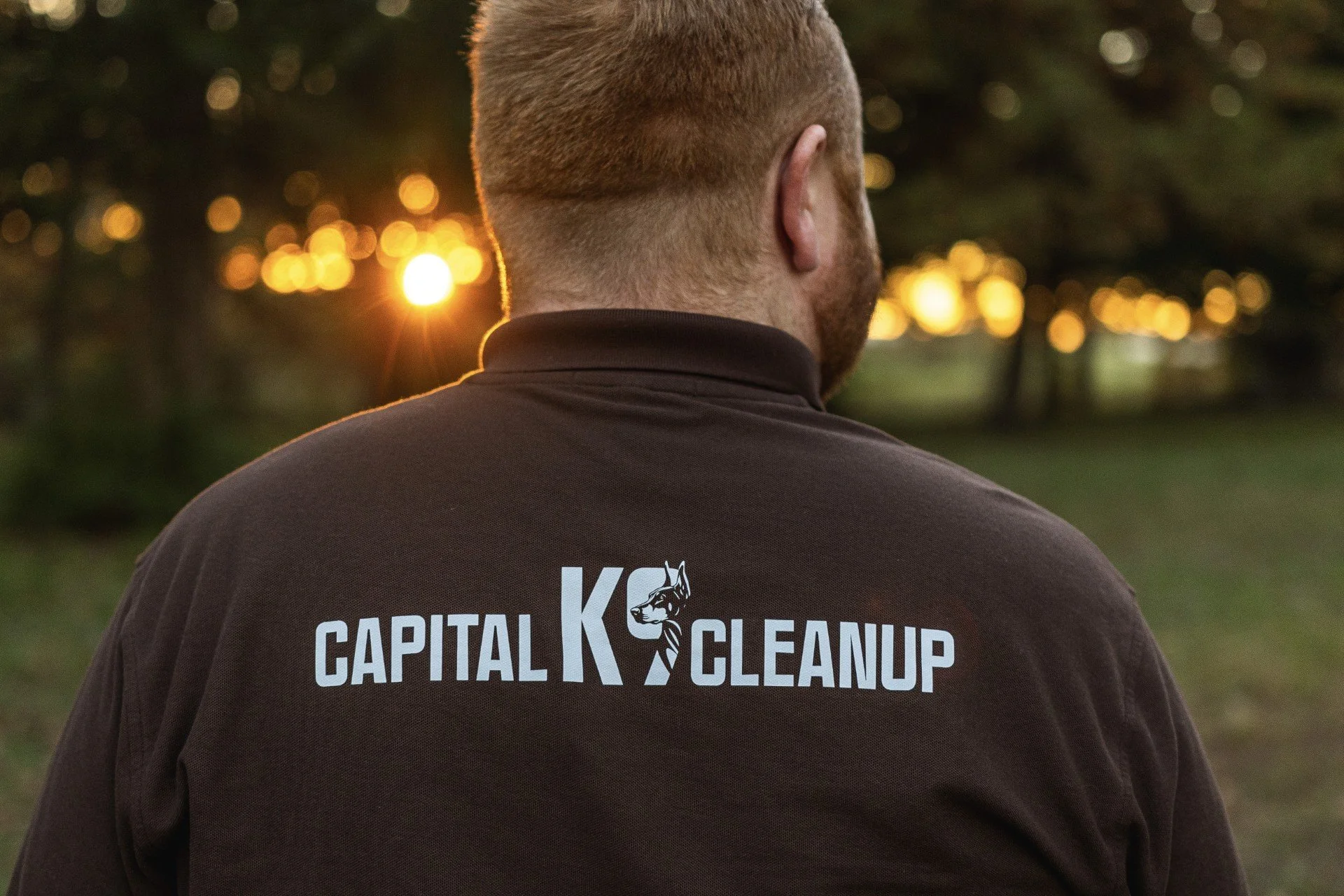 Back of a man wearing a dark gray t-shirt with white text that reads "Capital K Cleanup" and a dog logo. The man has short, light brown hair and is outdoors during sunset, with blurred trees and warm sunlight in the background.