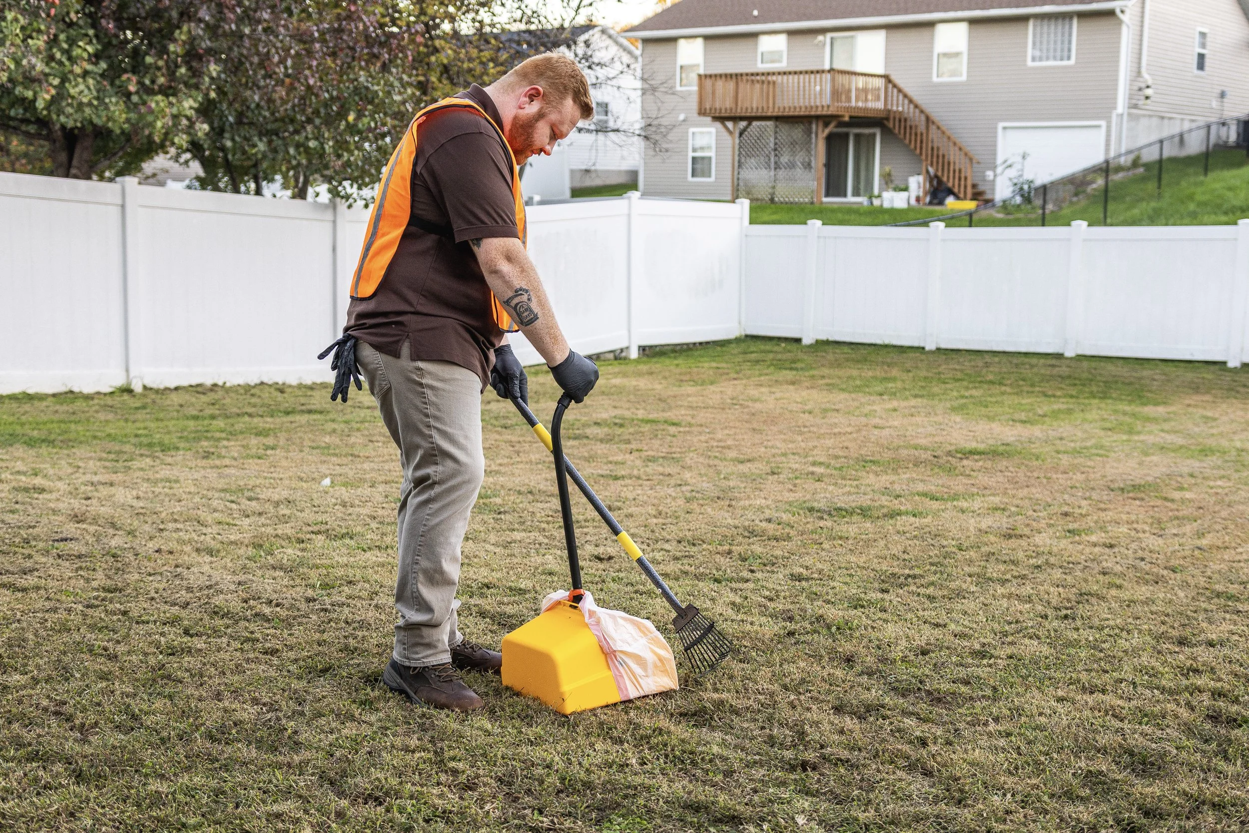 A man in work clothes and safety vest is using a metal detector on a grassy backyard, with a yellow paint sprayer and a dustpan nearby. There is a white fence and a residential house with a deck in the background.
