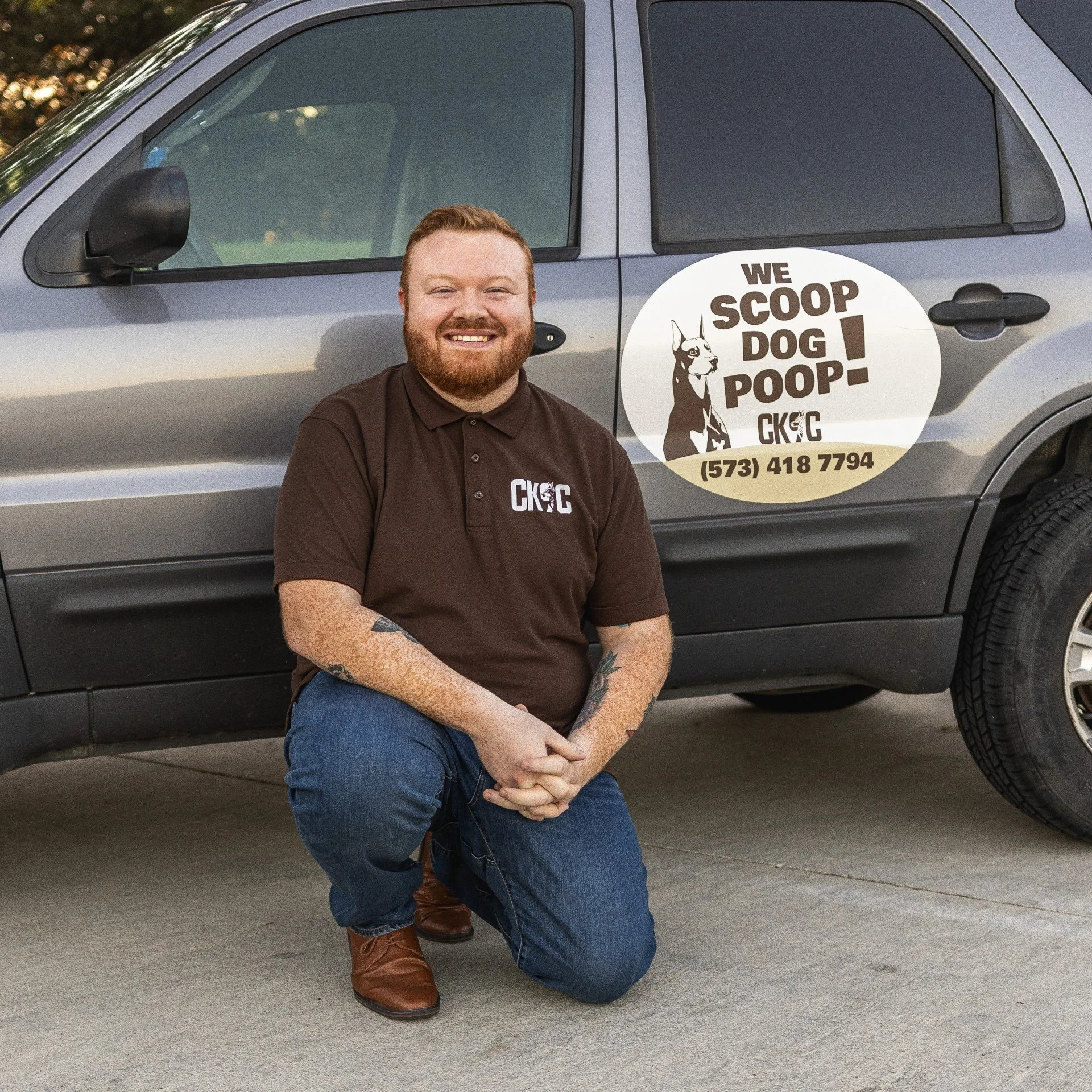 Man kneeling in front of a gray vehicle with a sign that reads "We scoop dog poop!" and a phone number, wearing a brown polo shirt and jeans.