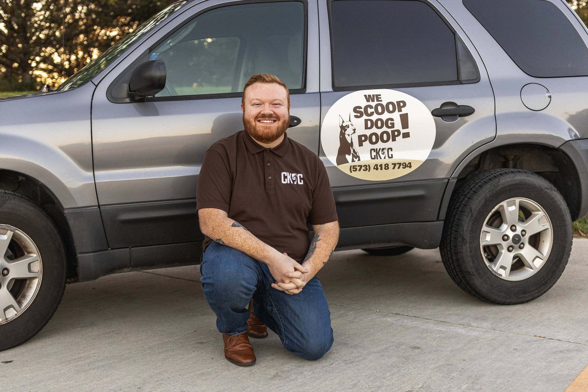 A man with red hair, beard, and tattoos kneeling on one knee in front of a gray vehicle with a decal on the side. The decal features a dog and the text 'We scoop dog poop' along with a phone number. The man is wearing a brown polo shirt with a logo and blue jeans, and is smiling at the camera.