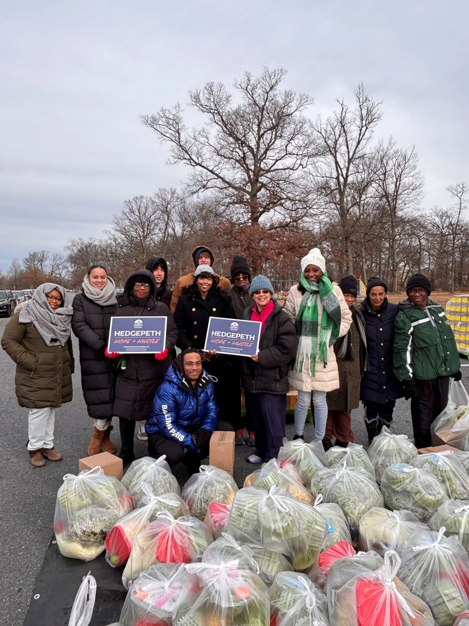 Volunteers holding Hope + Hustle signs after packing meals and standing in front of packed bags in front of some trees