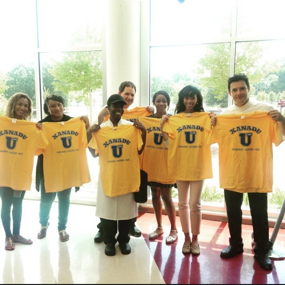 Group of eight ethnically diverse people holding yellow T-shirts with 'KANADU U' printed on them, standing indoors near large window with trees outside.