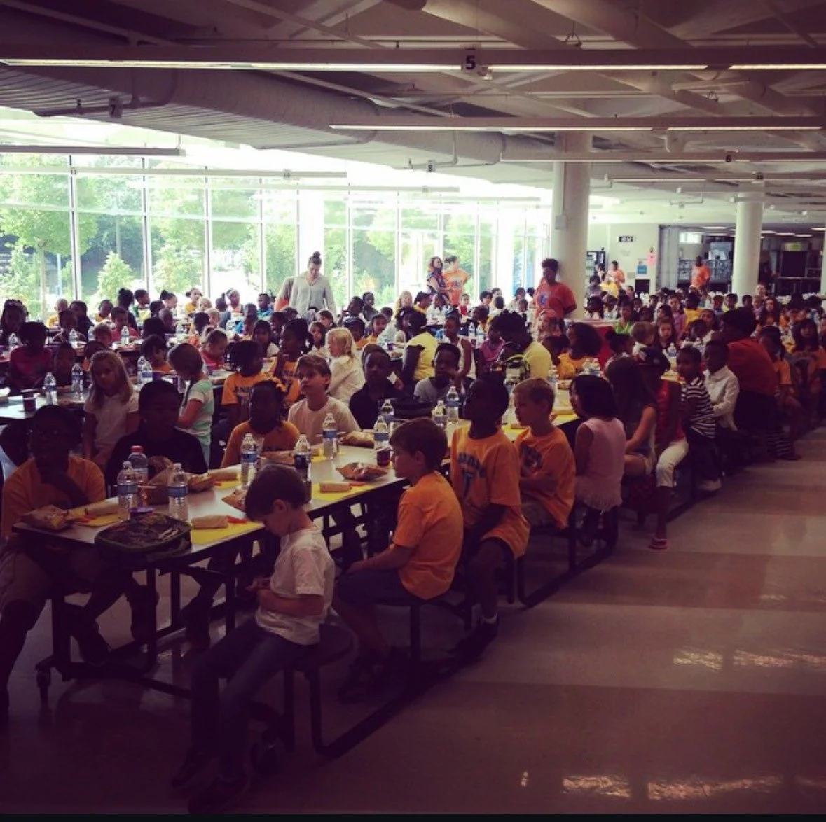 A large group of children seated at tables in a brightly lit cafeteria, some wearing orange shirts, with bottled water and meal trays in front of them, and adults walking around overseeing