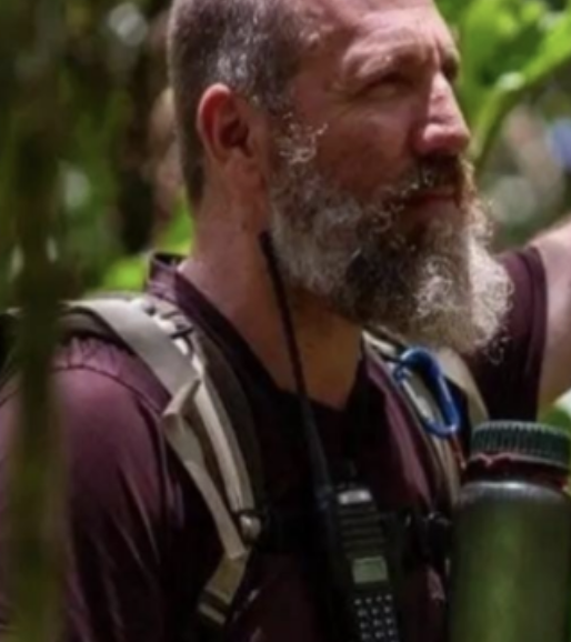 Homem com barba grisalha, usando mochila, em contato com a natureza, com vegetação ao fundo.