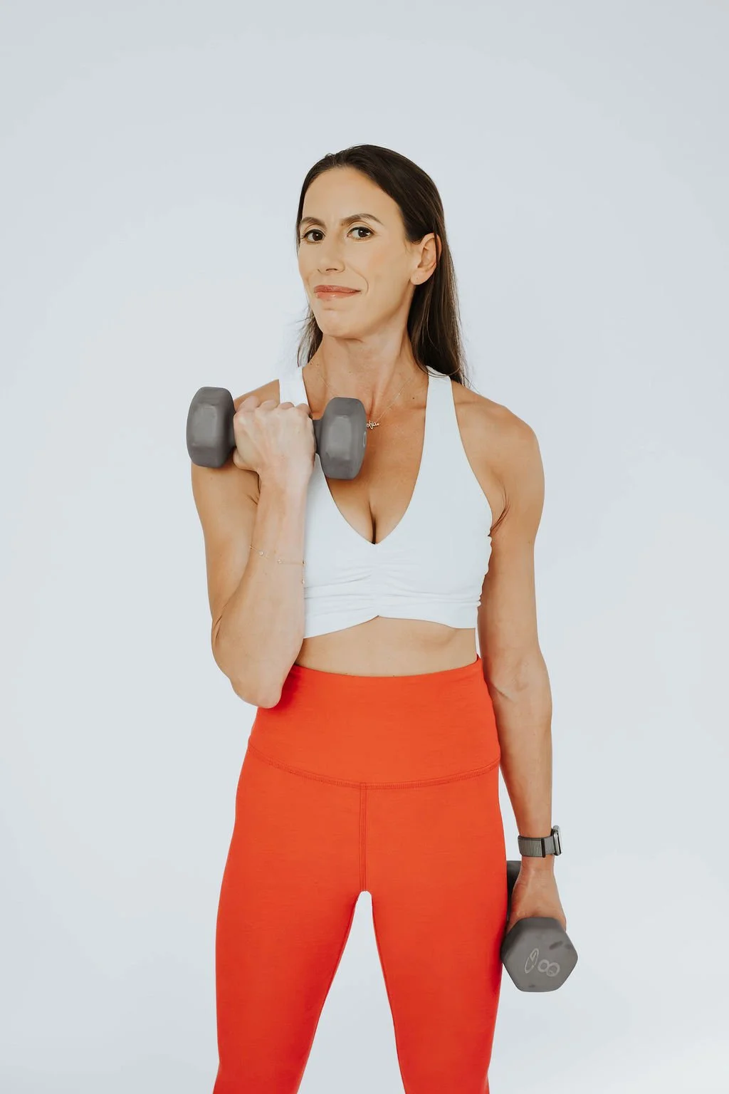 A woman in athletic clothing holding dumbbells, lifting one with her right hand and the other in her left hand, standing against a plain light-colored background.