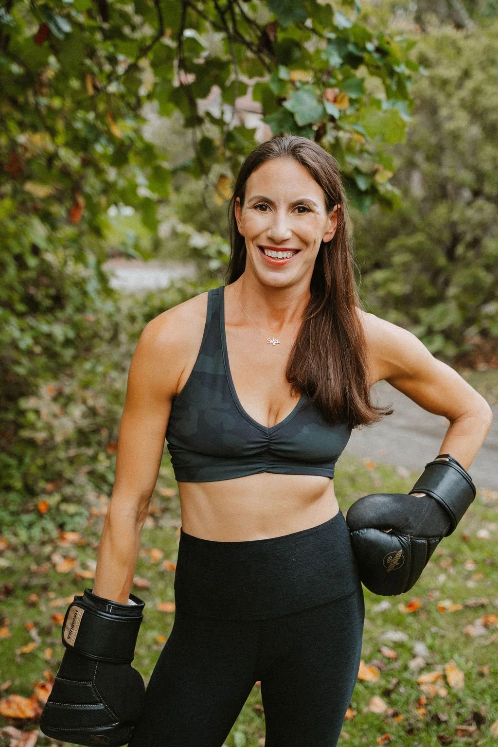 A woman outdoors wearing workout clothes and boxing gloves, smiling at the camera, surrounded by greenery.