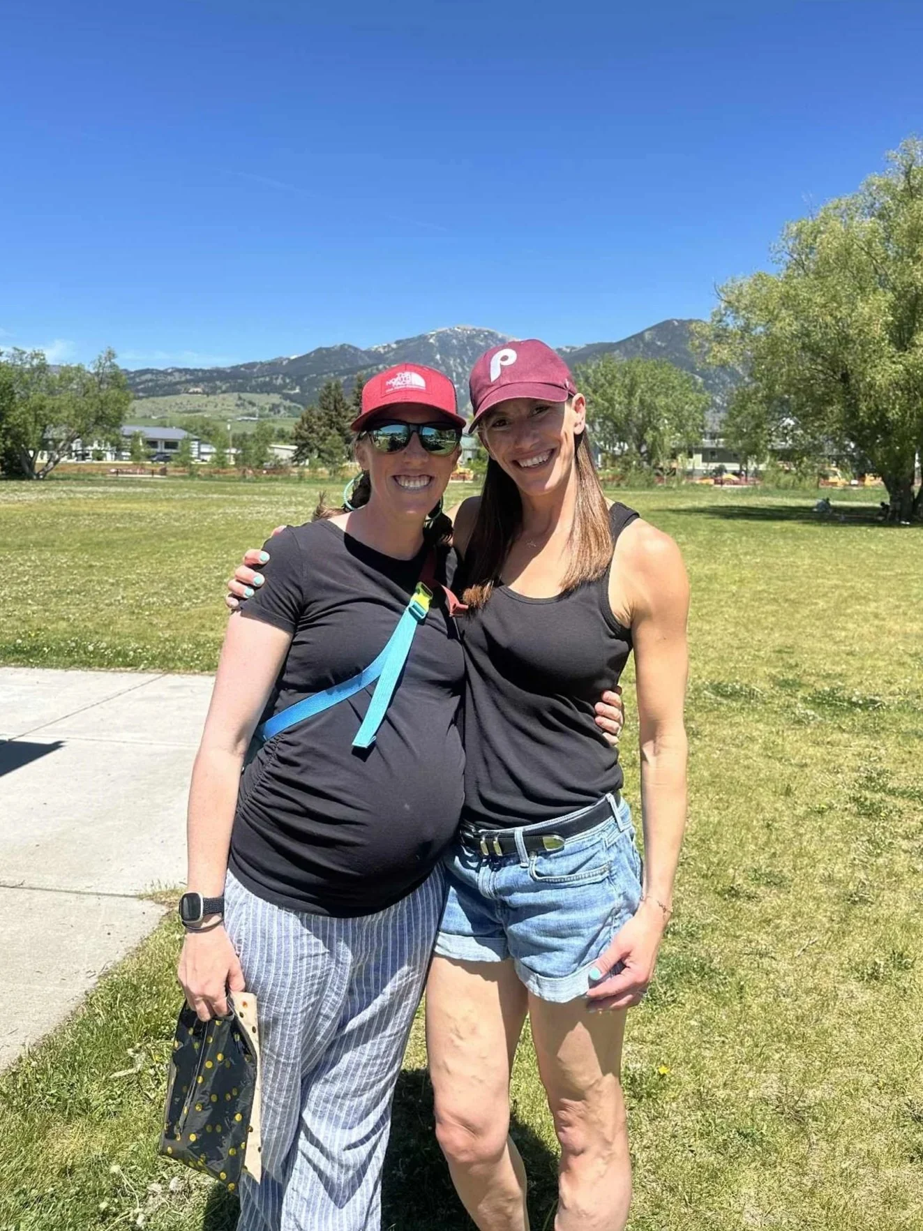 Two women standing outdoors on a grassy field, smiling with their arms around each other. One woman is pregnant, wearing sunglasses, a red cap, a black T-shirt, and striped pants. The other woman is wearing a maroon cap, a black tank top, and denim shorts. In the background, there are trees, houses, and mountains under a clear blue sky.