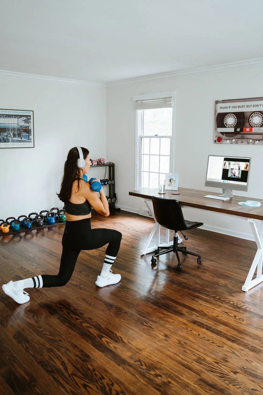 A woman exercising with dumbbells in a home office or exercise space, with kettlebells and a computer on a desk, a window providing natural light, and framed artwork on the walls.