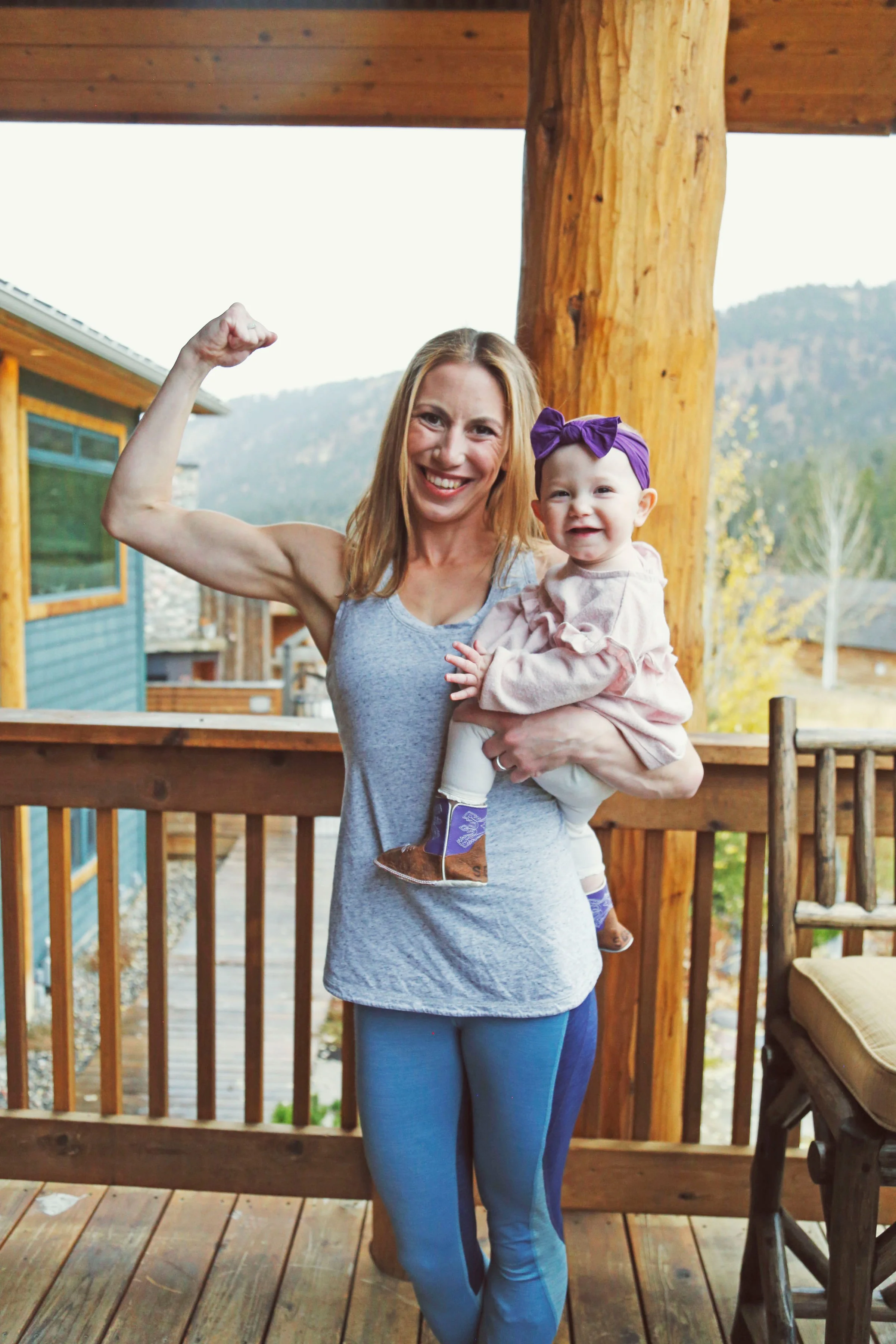 A woman flexing her arm muscle while holding a smiling baby girl on a wooden balcony outdoors.